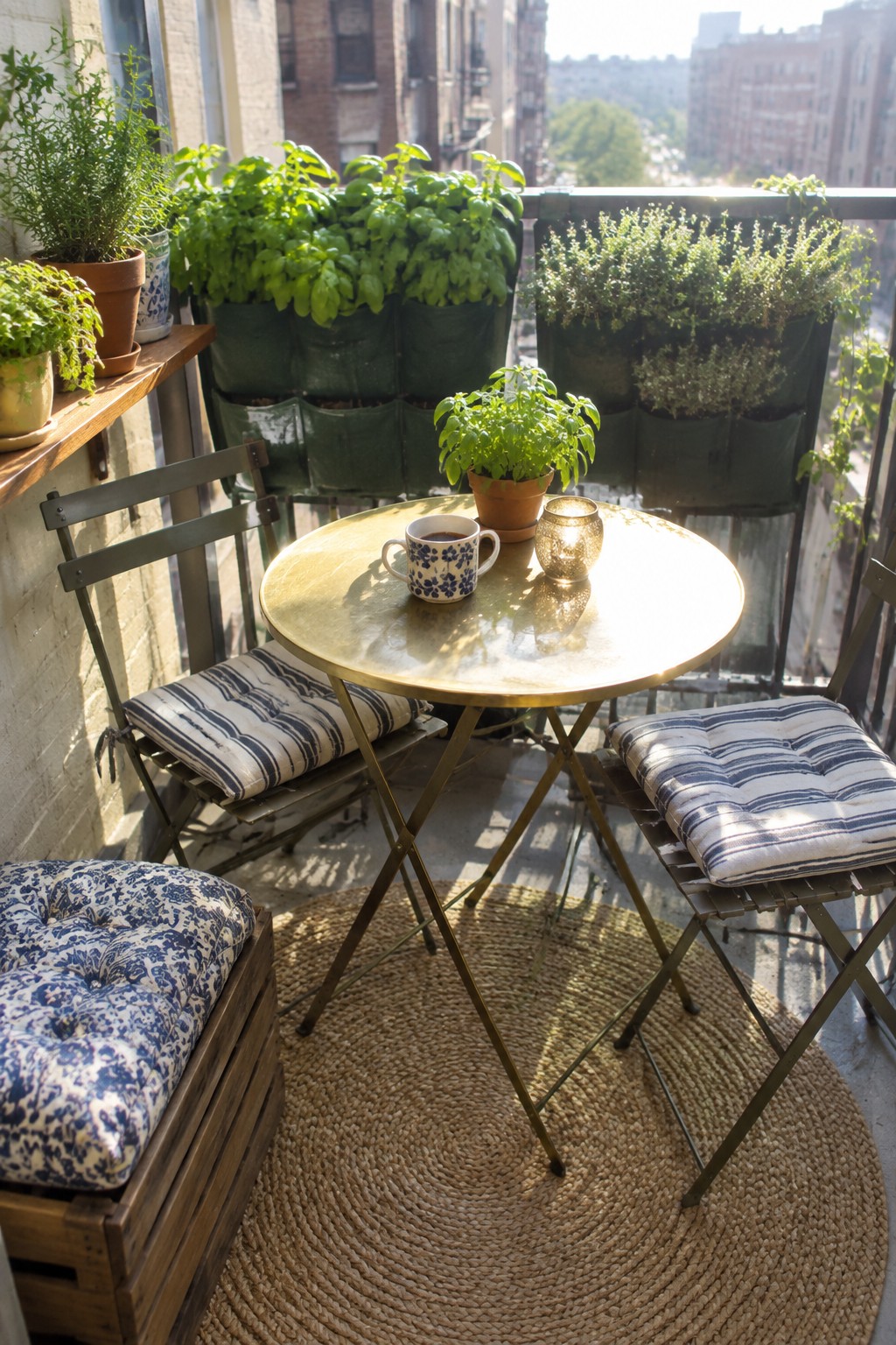 A round brass table with two metal folding chairs sits on a woven rug on a city balcony, surrounded by potted herbs on the railing and a wooden crate stool.