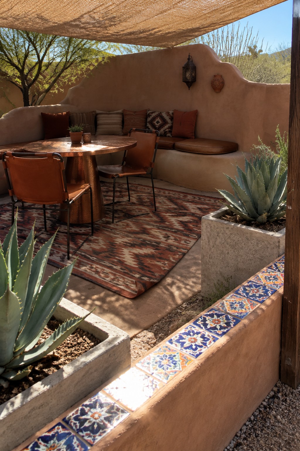 Outdoor patio with round metal dining table, leather chairs, patterned area rug, built-in stucco bench with cushions, and concrete planters with agave plants under a shade sail.