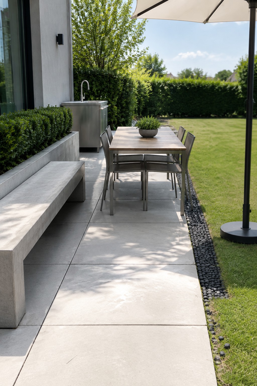 A rectangular concrete paver patio extending from a house, featuring an outdoor dining table with chairs, a long concrete bench, an outdoor sink, and a pebble border separating the pavers from the lawn.