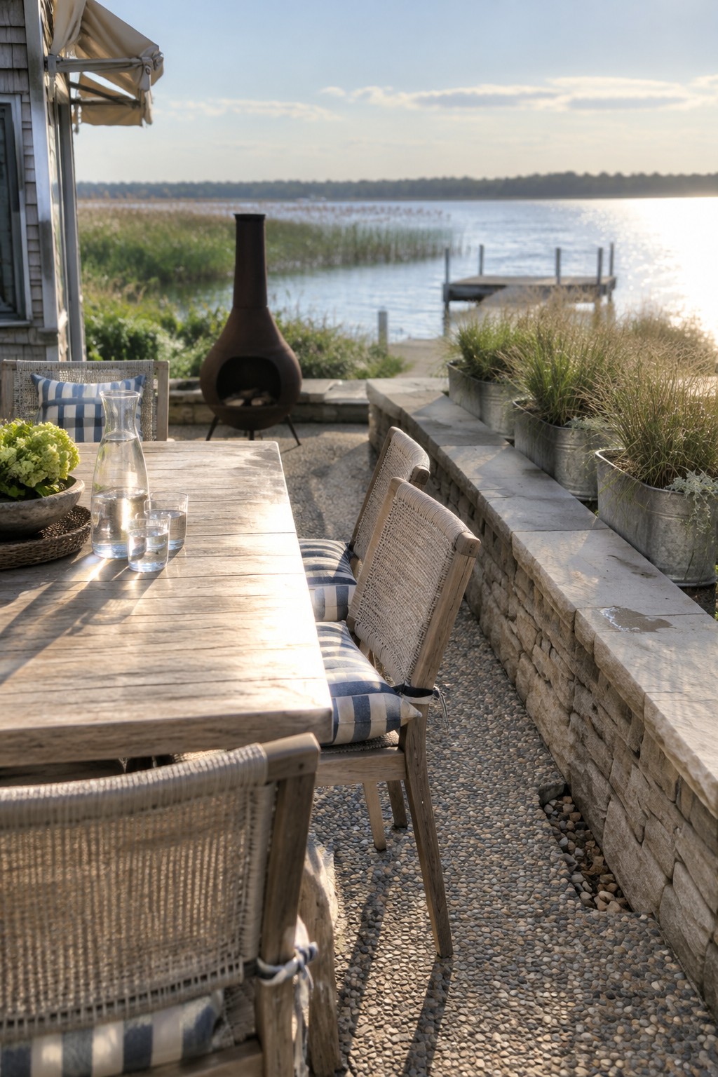 Sunlit wooden table with blue-striped chairs overlooking lake, dock, and chiminea