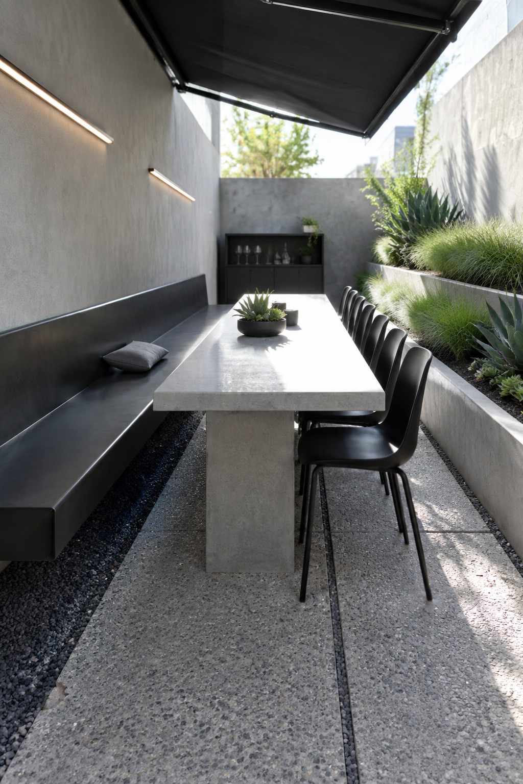 A narrow outdoor patio with a long concrete dining table, black bench on the left, black chairs on the right, and raised planters with grasses and succulents along one wall.