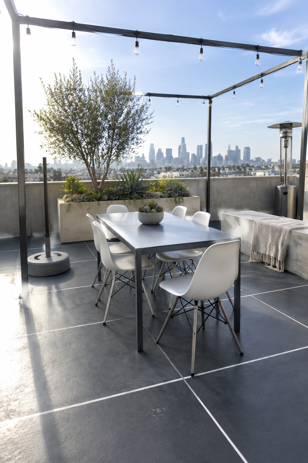 A rooftop patio features a dark gray painted concrete floor, a rectangular dining table with white chairs, a concrete planter with a small tree, string lights on a metal frame, and a city skyline in the background.