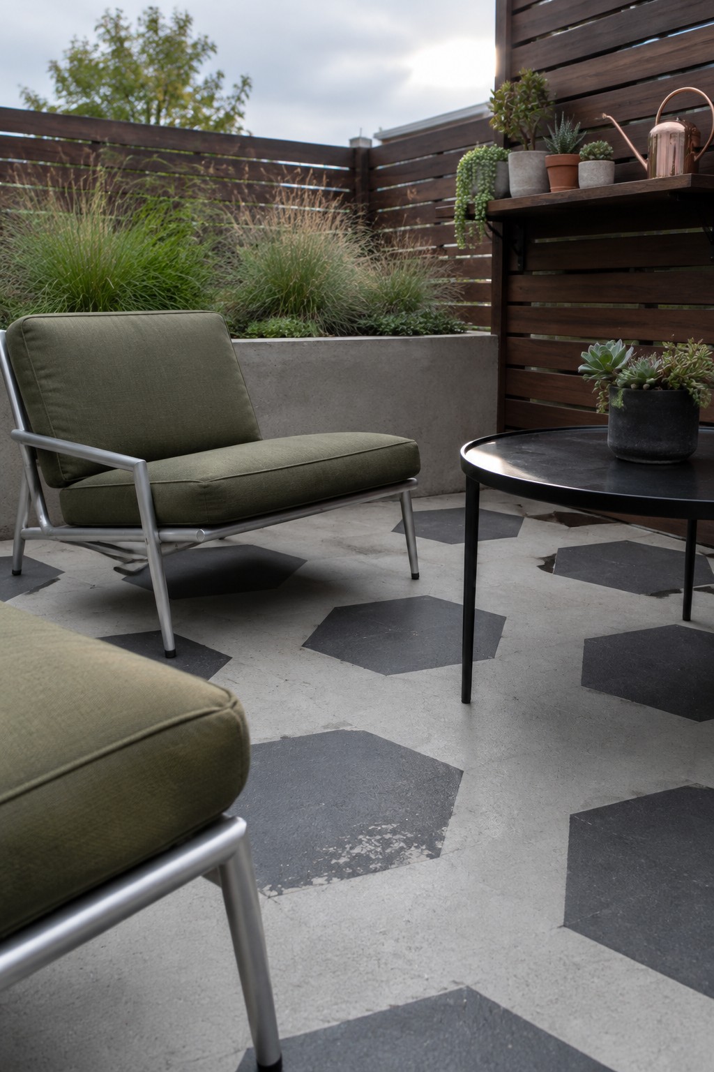 An outdoor patio with green upholstered lounge chairs, a round black table, potted plants on a wooden shelf, and a concrete floor painted with dark hexagonal shapes.