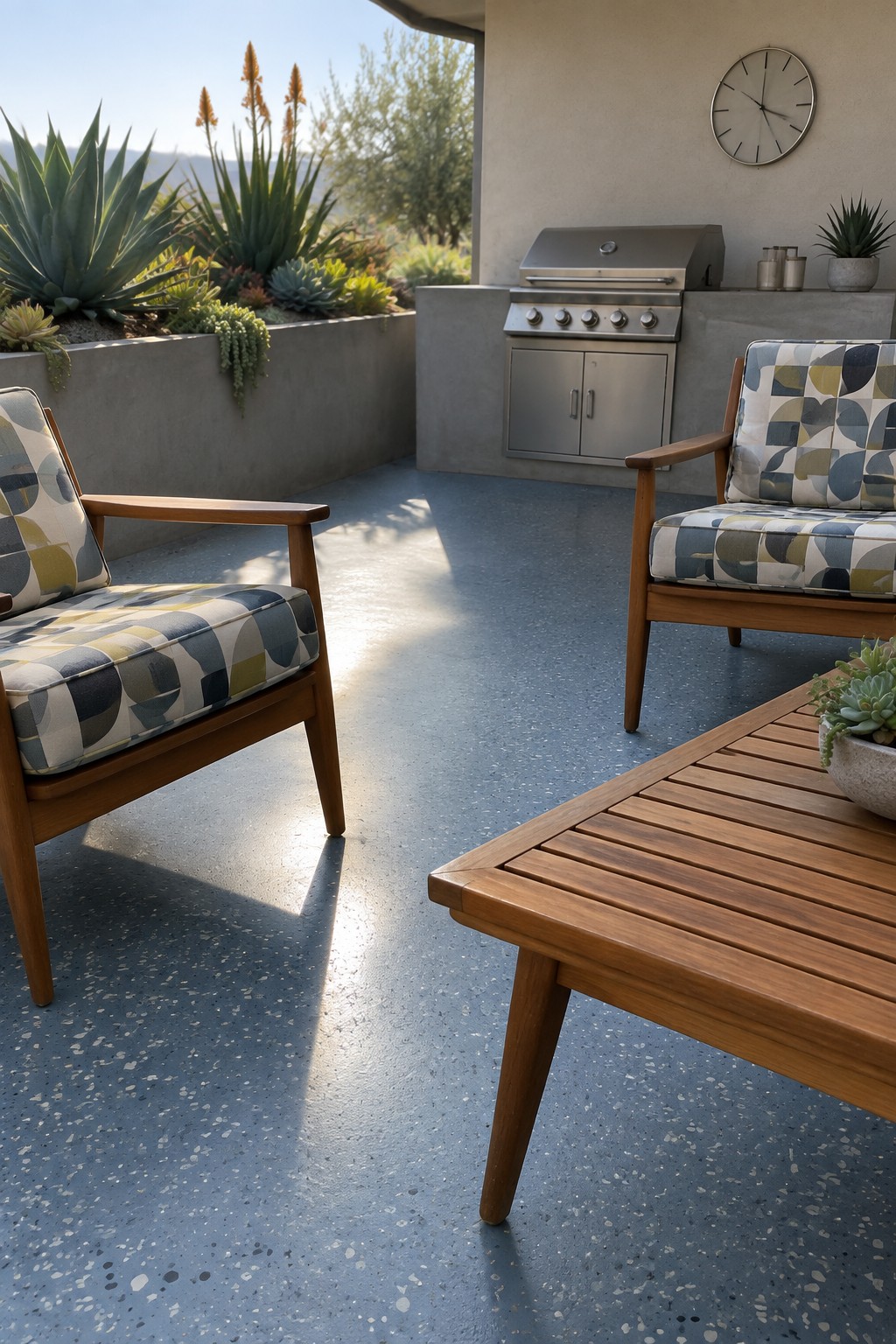 Two wooden patio chairs with geometric cushions face each other across a slatted coffee table on a blue speckled concrete floor, with a built-in stainless steel grill along the back wall.