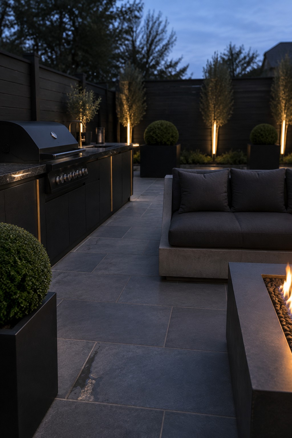 A modern outdoor patio at dusk featuring a long black grill island with a built-in grill on the left, a dark sectional sofa facing it, a rectangular fire pit in the foreground, and several tall potted trees with vertical lighting along a dark fence.