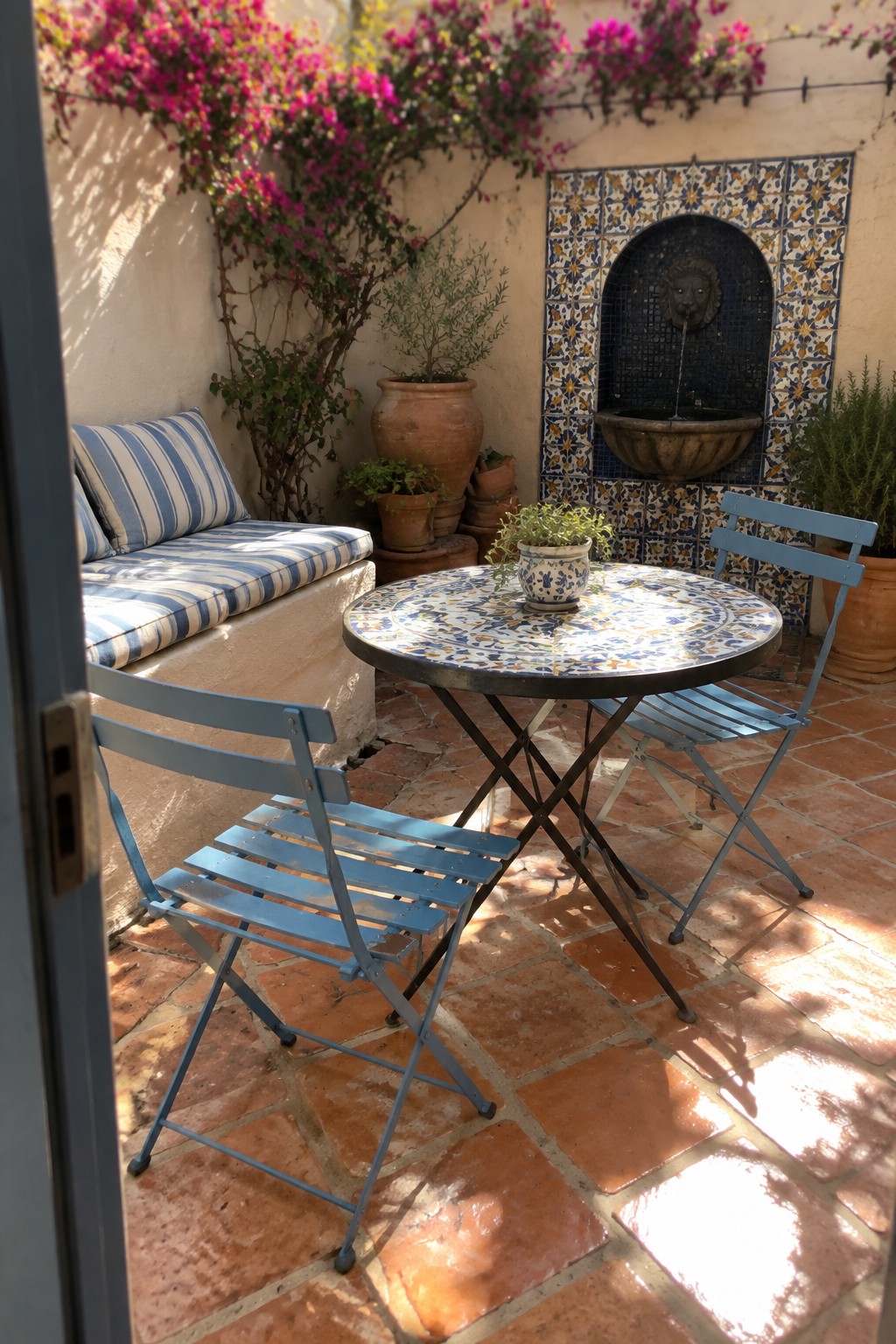 A small patio with terracotta tiles, a round mosaic table, two blue folding metal chairs, a built-in cushioned bench, and a tiled fountain on the back wall.