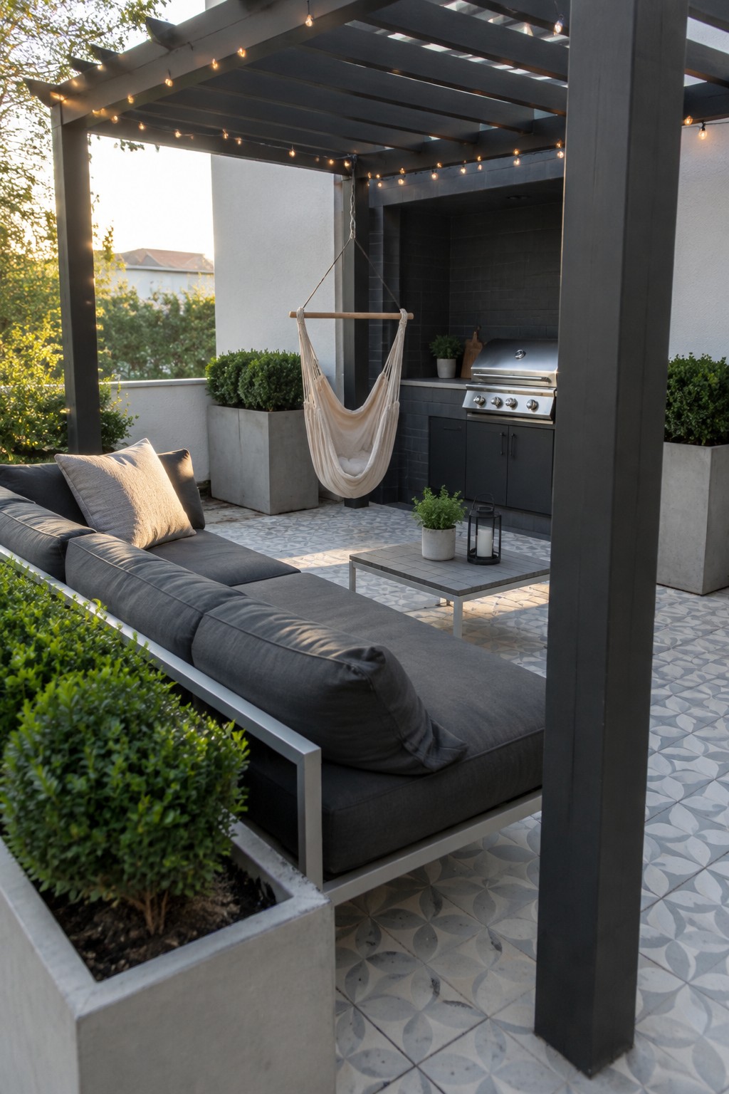 An outdoor patio under a dark pergola with solar string lights, featuring a gray sectional sofa, hanging hammock chair, coffee table, and outdoor kitchen area with potted plants.
