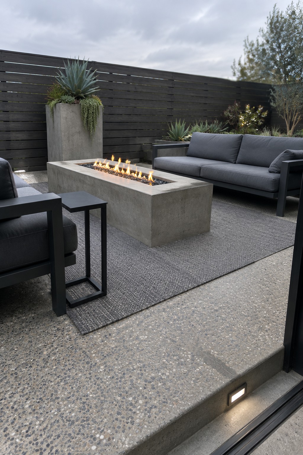 A rectangular concrete fire pit with active flames rests on a large gray woven rug on a patio, with dark outdoor sofas arranged around it.