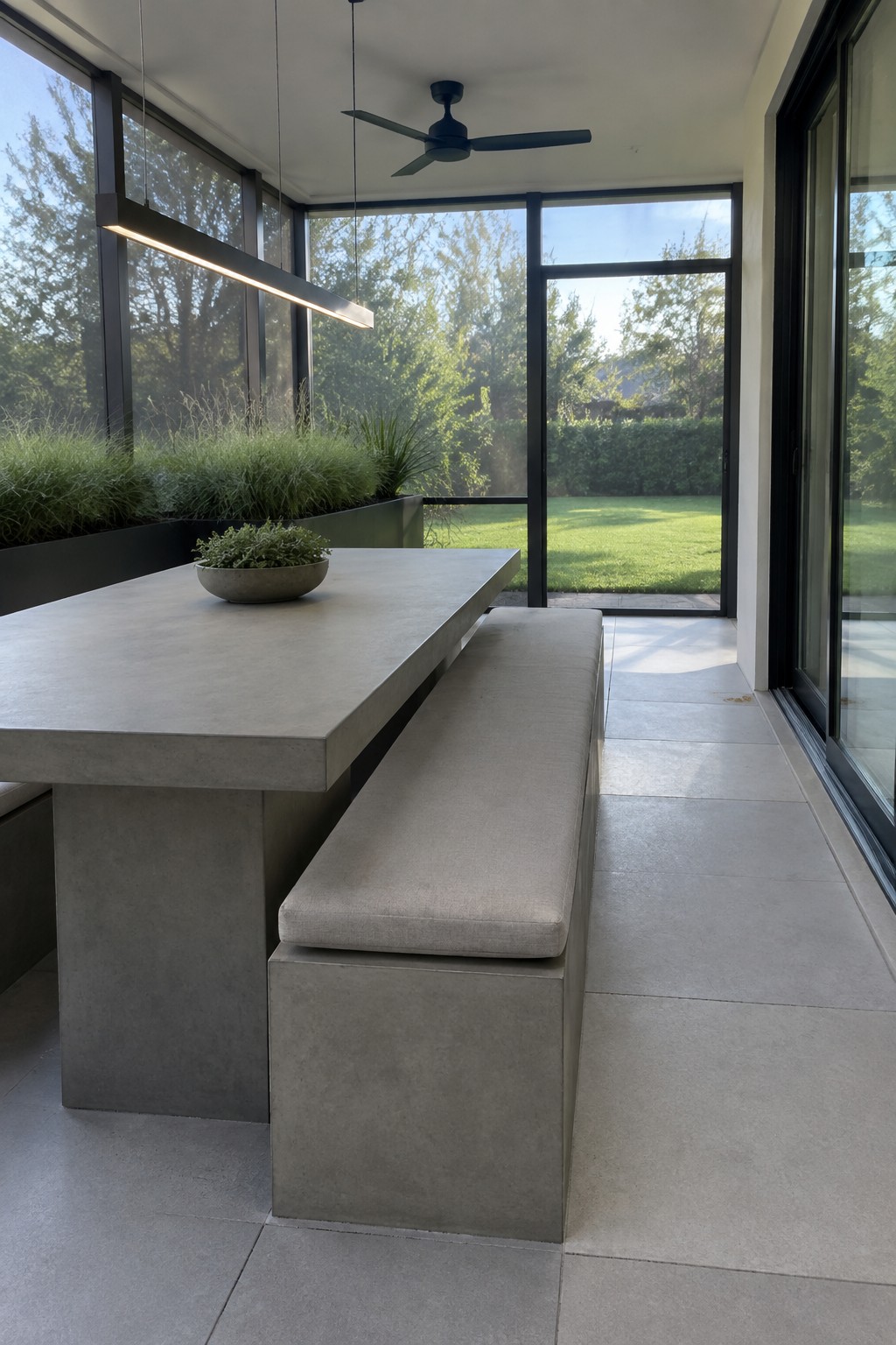 A long gray concrete dining table with a cushioned bench sits inside a screened patio with large glass doors opening to a lawn.
