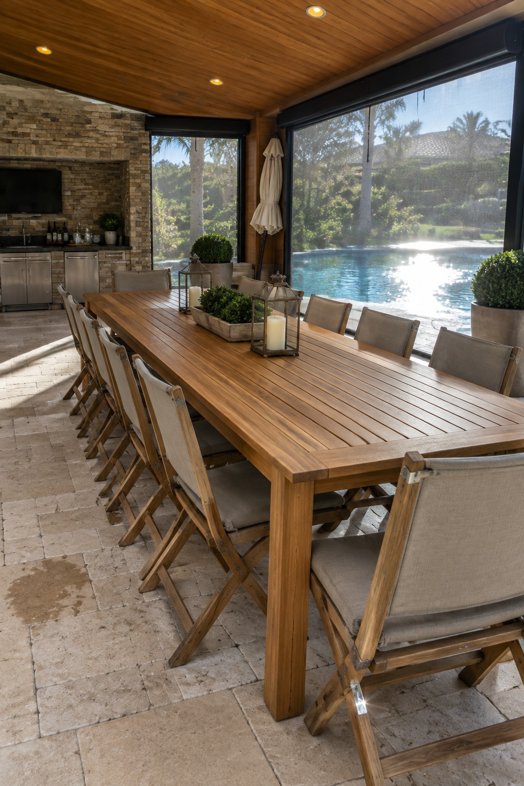 A long wooden dining table with multiple folding chairs arranged inside a screened patio overlooking a pool.