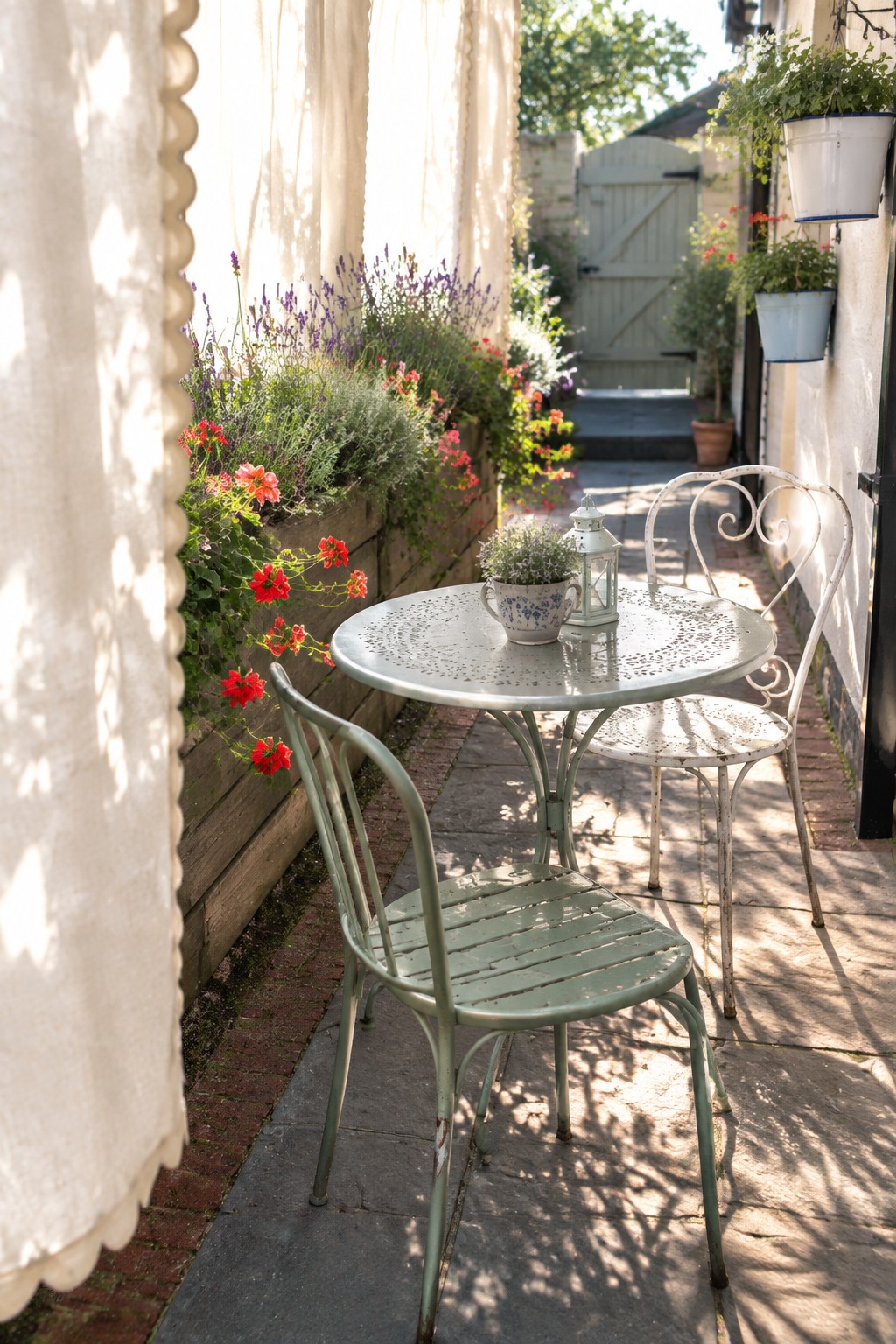 A small patio with a round metal table and two chairs next to a wooden planter box filled with red flowers and lavender, with a white scalloped curtain hanging on the left side.