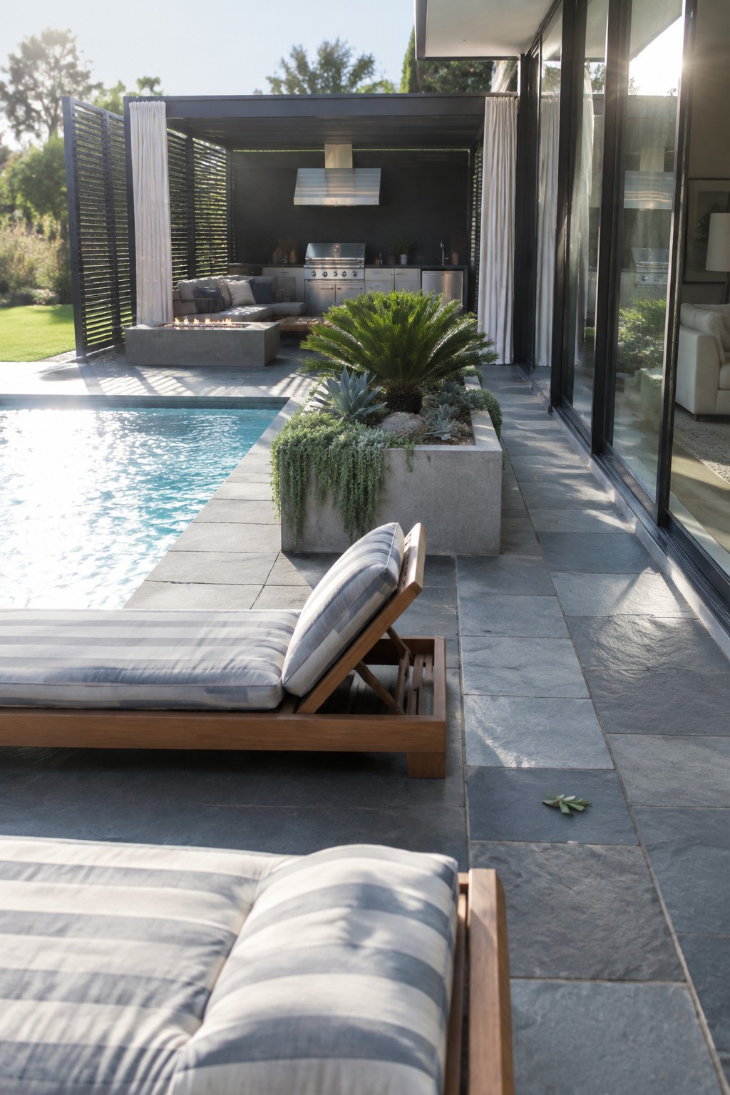 An outdoor patio area with a pool, wooden lounge chairs on stone tiles, a large concrete planter filled with plants, white curtains, a slatted screen, and an outdoor kitchen in the background.