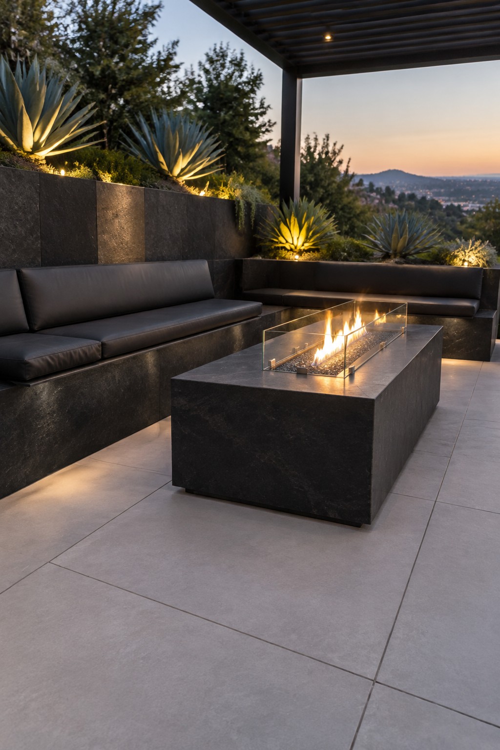 An outdoor patio with dark stone retaining walls forming L-shaped built-in seating around a rectangular fire pit, surrounded by agave plants and overlooking a hillside at dusk.