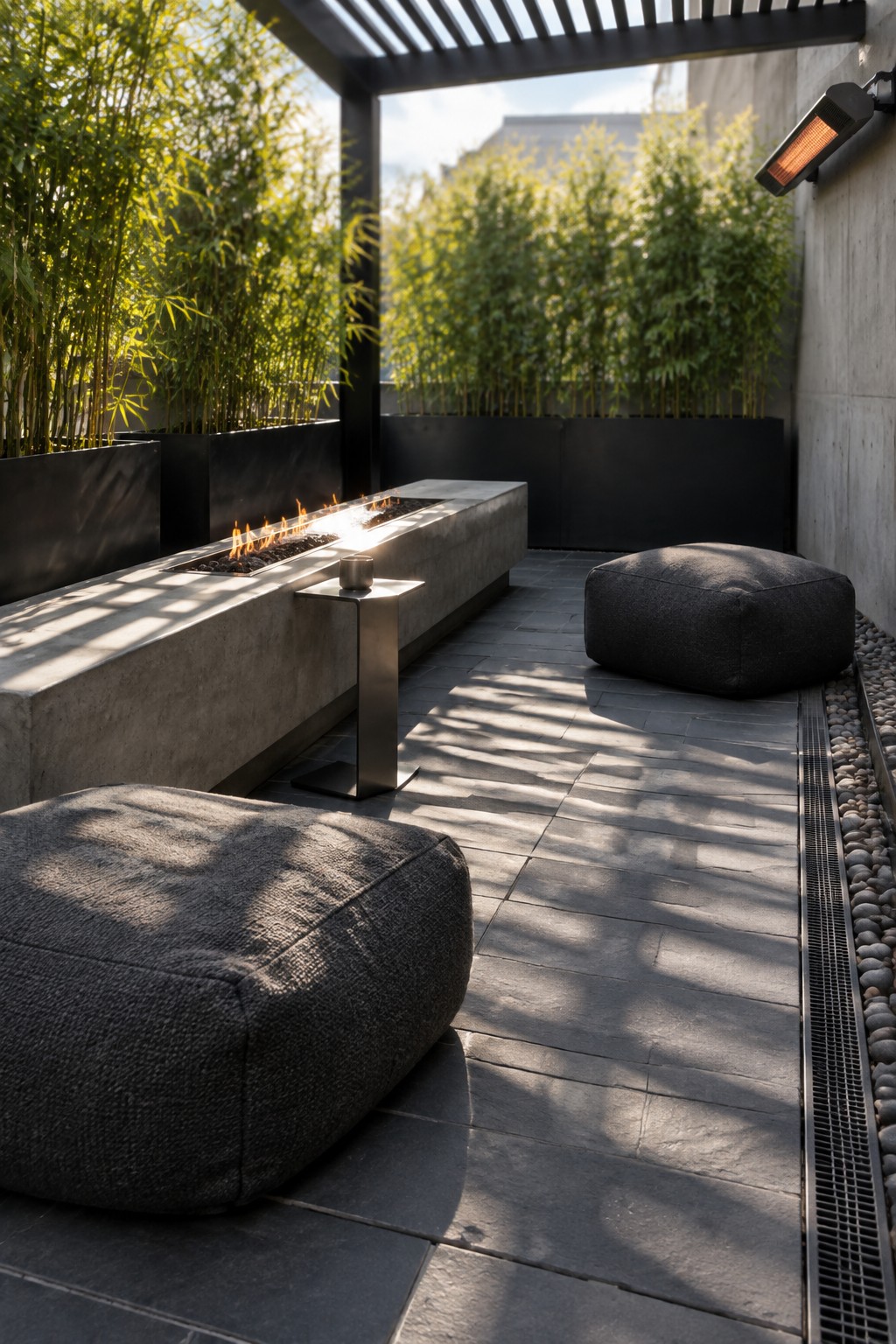 A small modern patio with a long concrete fire pit, two dark fabric poufs, a metal side table, and tall bamboo planters along a dark wall.