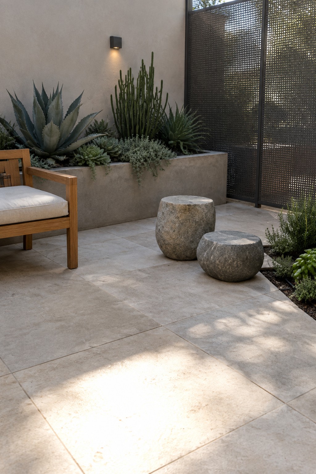 A small tiled patio with a wooden armchair, two round stone stools, and a long concrete planter filled with agave, cacti, and succulents next to a perforated metal screen.