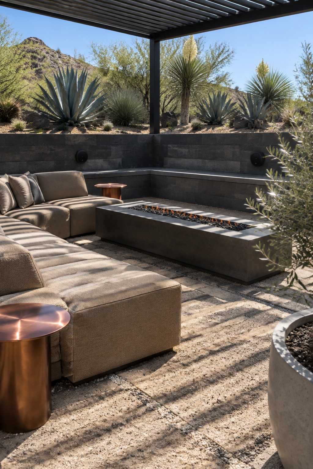 A large L-shaped modular sectional in beige fabric sits on a stone-paved patio around a rectangular concrete fire pit with active flames, under a dark metal pergola in a desert setting with agave plants and mountains visible.