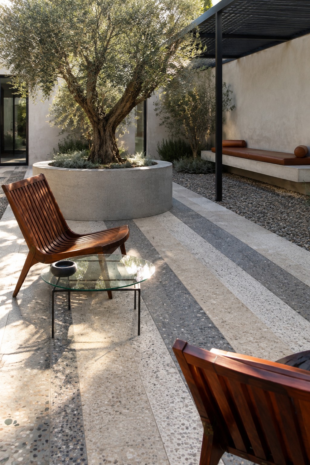 An outdoor patio with striped geometric pavers in neutral tones, wooden lounge chairs, a round glass table, and a large concrete planter holding an olive tree.