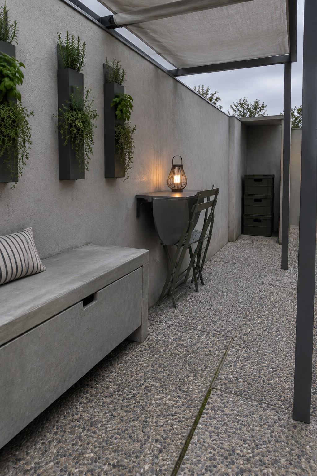 A narrow outdoor patio with a concrete wall, built-in bench, vertical planters, wall-mounted drop-leaf table with two folding chairs, and a lit lantern.