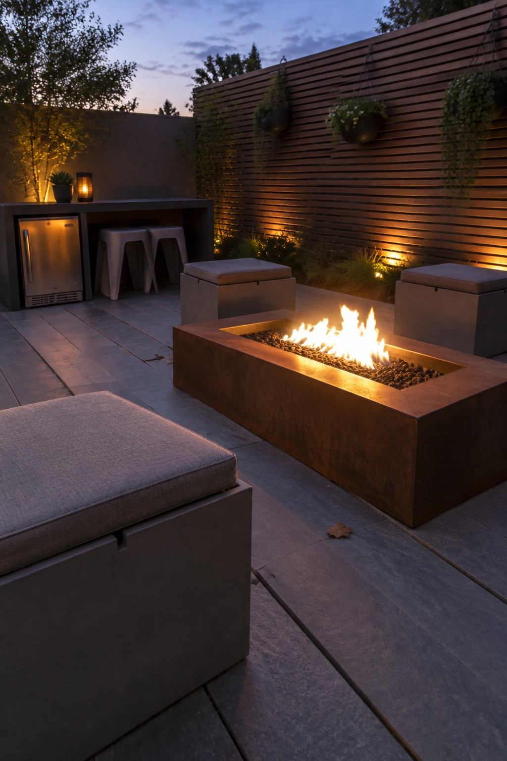 A rectangular metal fire pit with active flames sits on a wooden deck at dusk, surrounded by gray cube ottomans with fabric cushions, next to a built-in bar with a stainless steel refrigerator and metal stools.