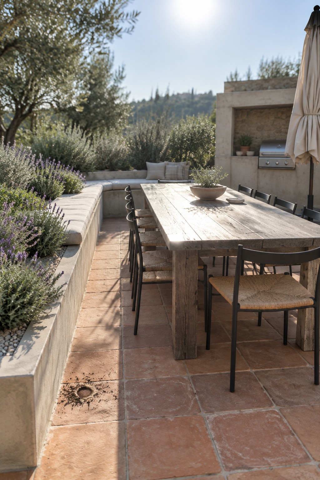 A long wooden dining table with black chairs sits on a terracotta tile patio next to a concrete planter filled with lavender and a cushioned built-in bench.