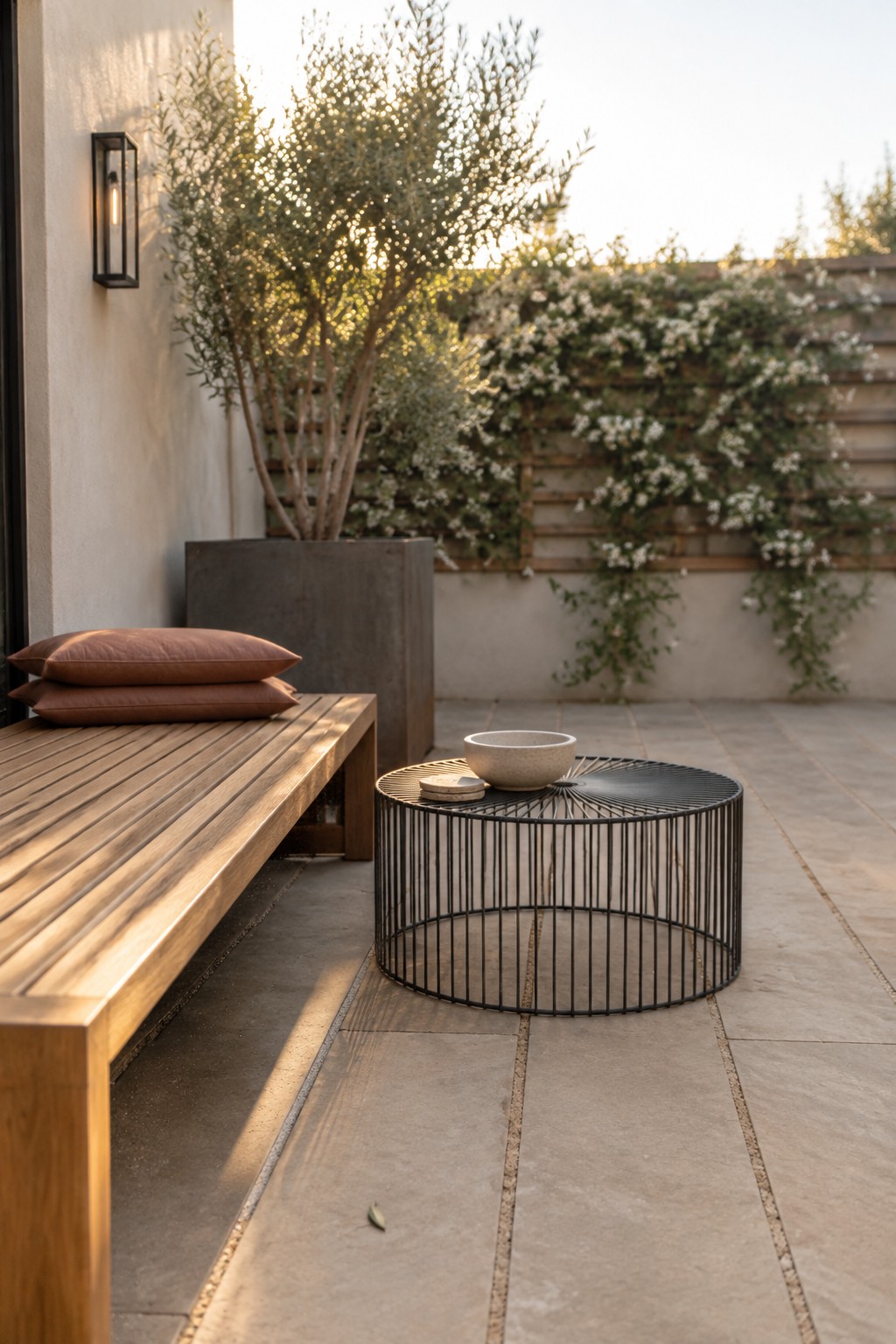 A wooden bench with two brown cushions next to a round black wire frame coffee table holding a ceramic bowl on a stone tiled patio, with a large potted tree and flowering vines on a trellis in the background.
