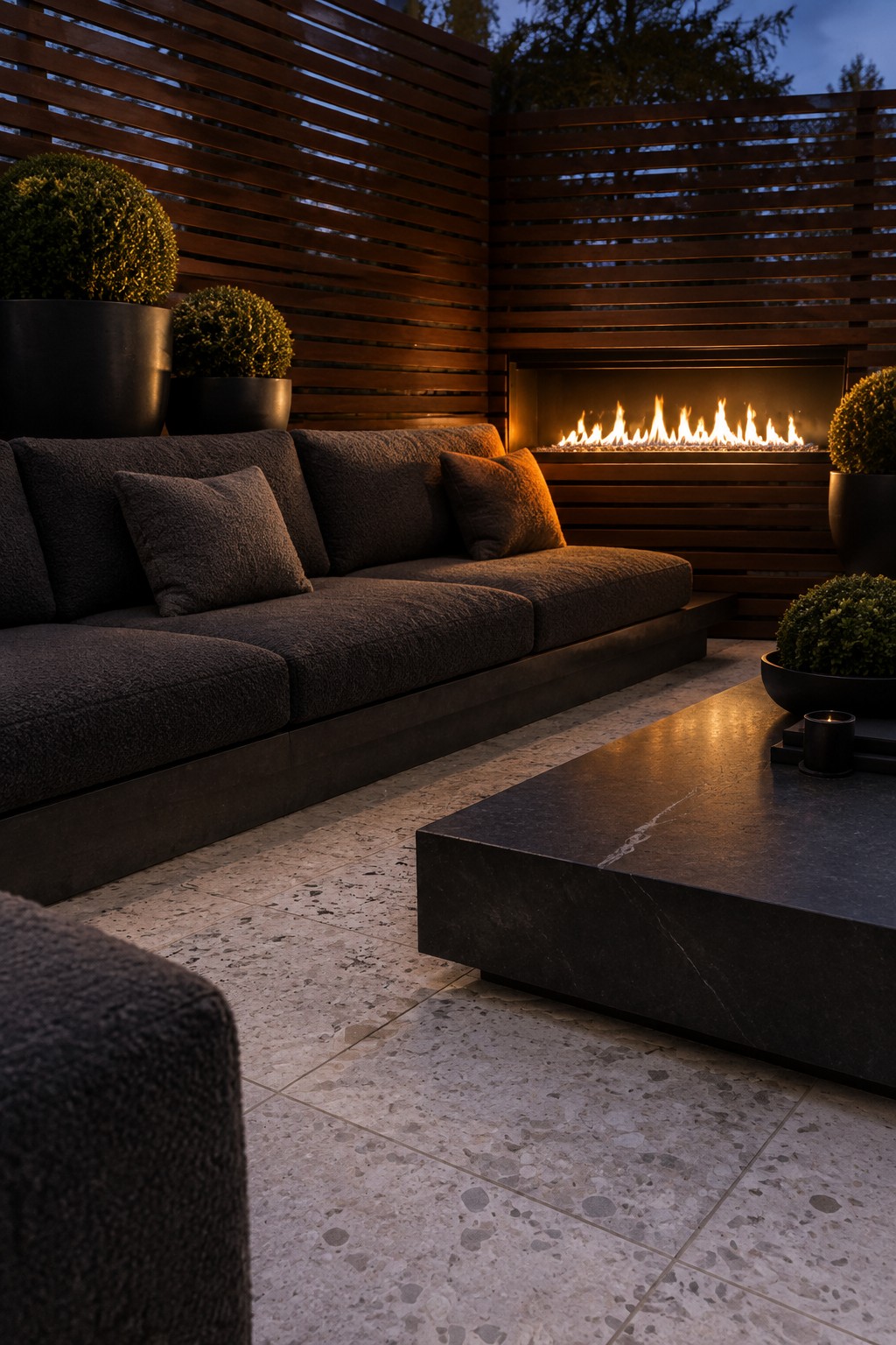 An outdoor patio enclosed by horizontal wooden slat screens features a dark sectional sofa, stone coffee table, built-in fire pit, and several potted shrubs.