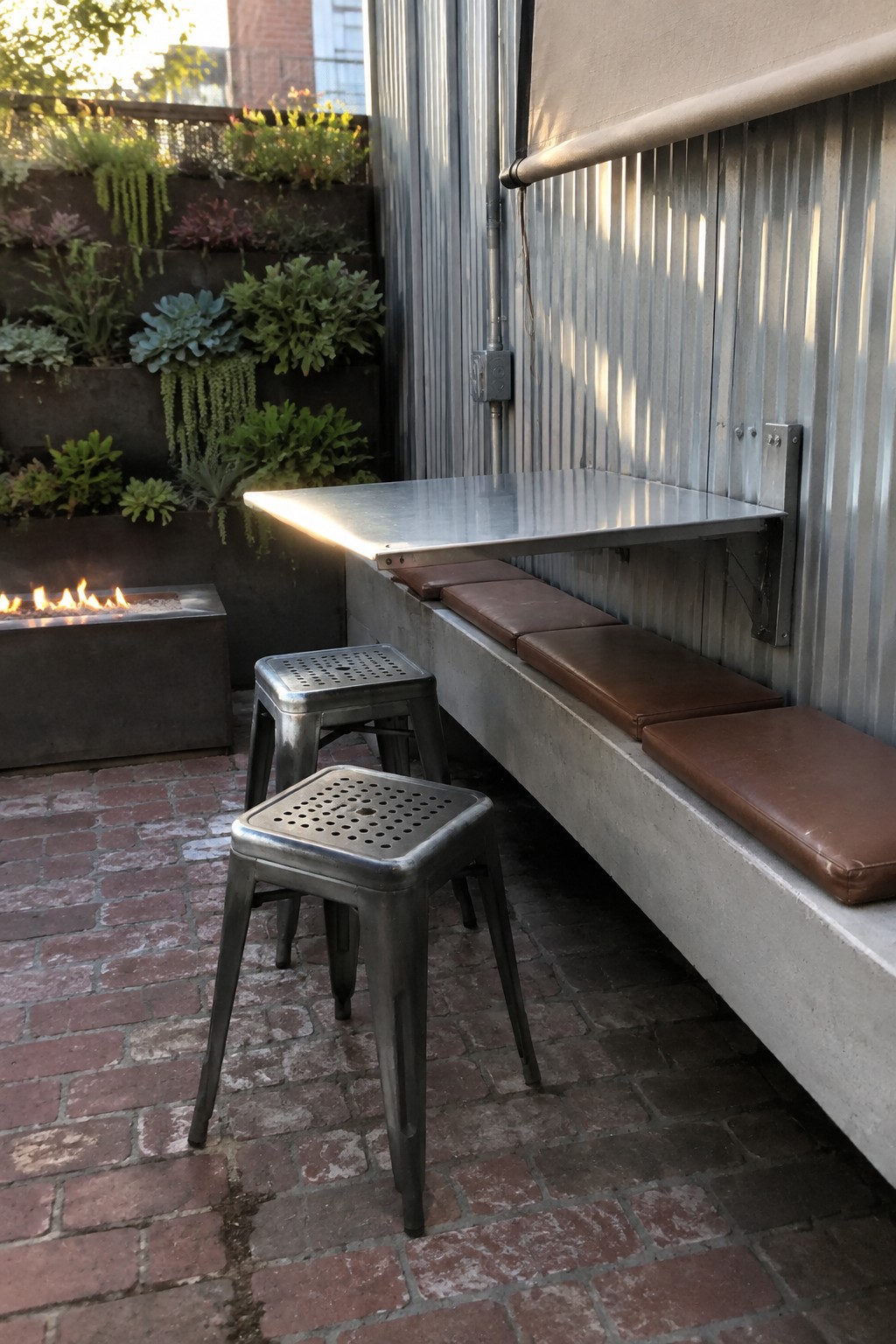A compact outdoor patio features a wall-mounted fold-down metal table, brown cushioned bench seating, two perforated metal stools on brick paving, and a raised garden bed with plants beside a fire pit.