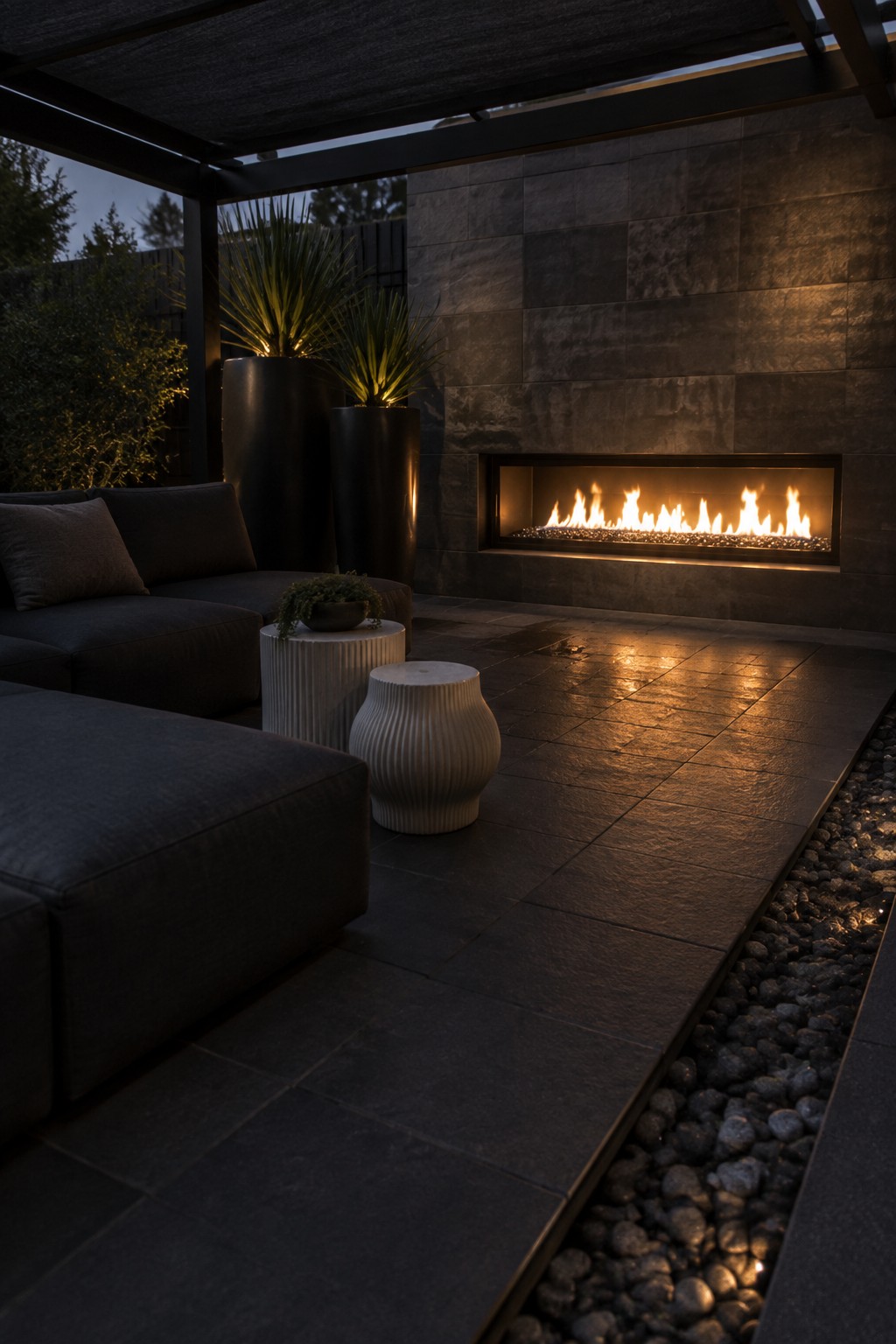 An outdoor patio at dusk featuring a long linear fireplace built into a dark stone wall, with a dark sectional sofa, two light ribbed side tables, and a strip of pebbles along the edge of the tiled floor.