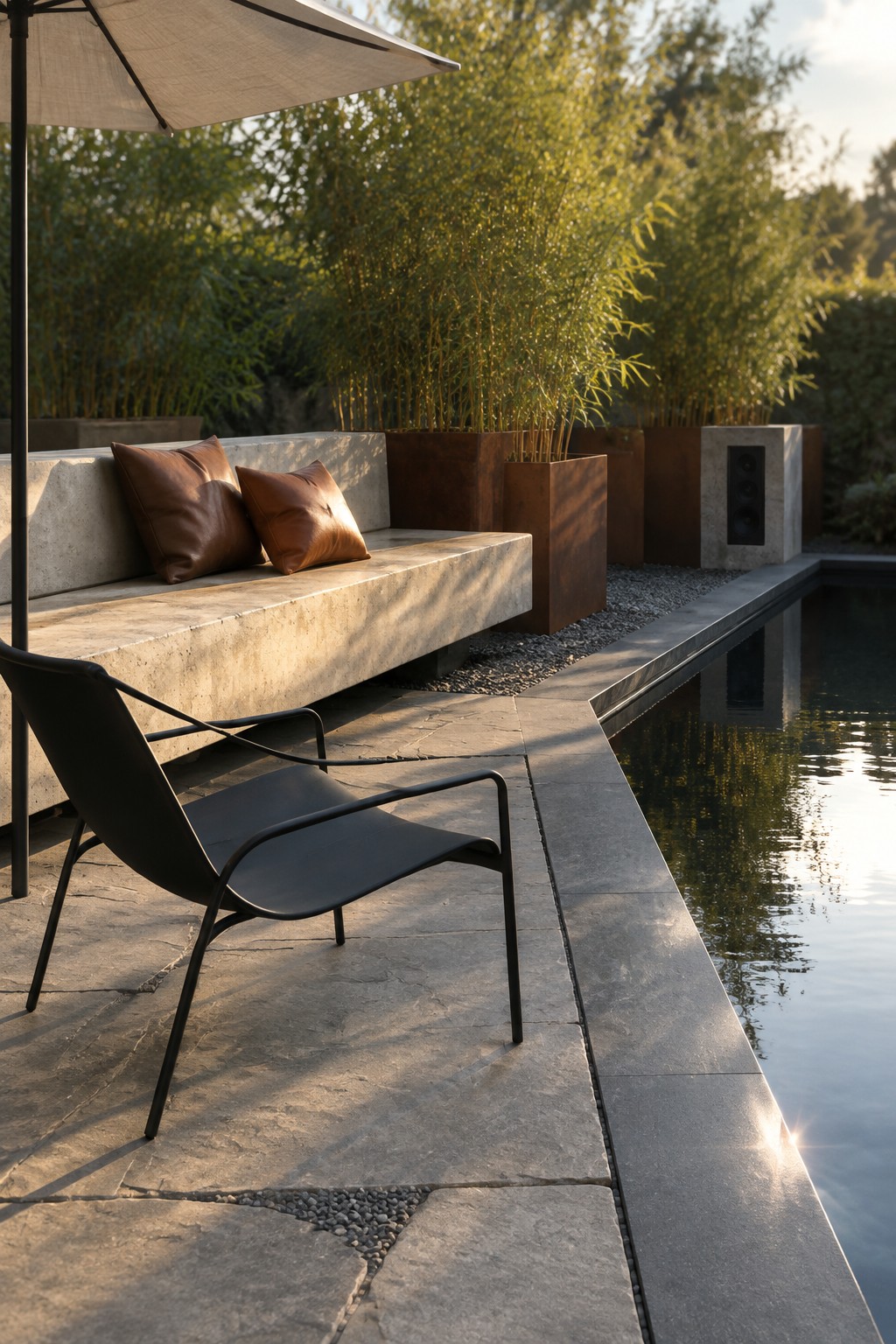 A modern poolside patio with a long concrete bench holding two brown leather cushions, a black metal lounge chair, a large beige umbrella, and tall planters with bamboo along the pool edge.