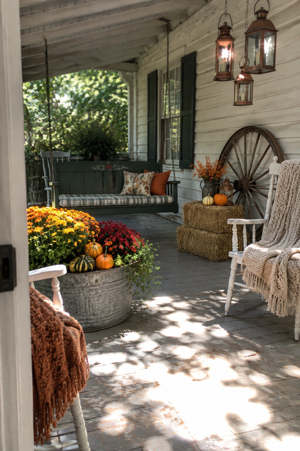 A white wooden porch with a green porch swing, a rocking chair covered by a beige knit throw, a metal tub filled with orange and red mums plus small pumpkins, and hay bales with lanterns against the house siding.