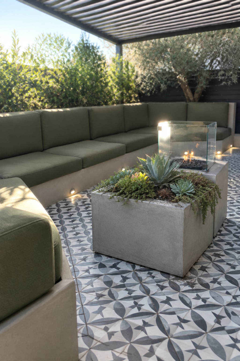 A concrete patio shows a large rectangular planter filled with succulents next to a glass fire pit, with green outdoor sofas arranged around it on patterned tile.