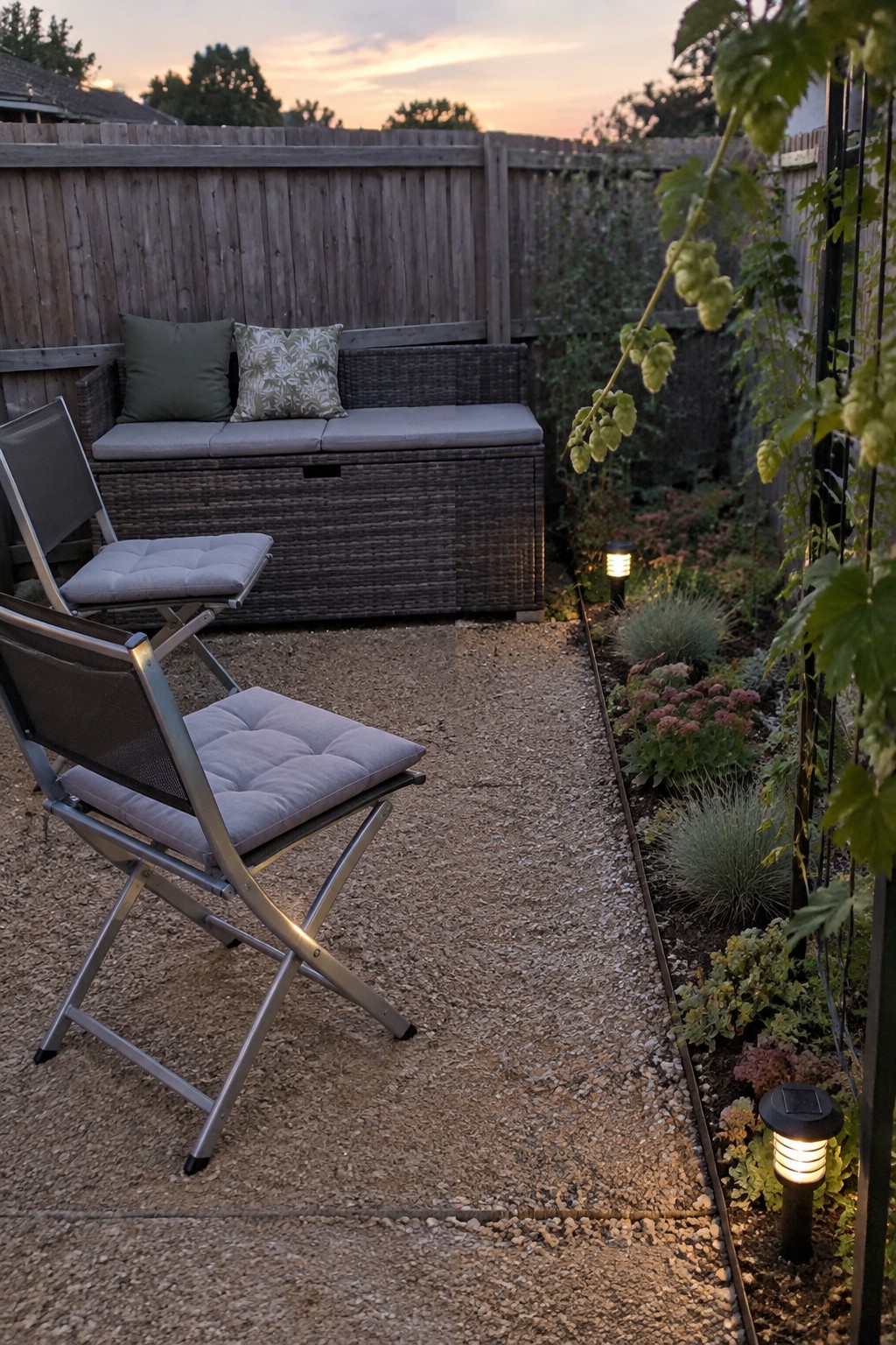 A gravel patio features a dark wicker storage bench with gray cushions against a wooden fence, two foldable metal chairs in the foreground, and a narrow garden bed with plants and solar lights along the right edge.