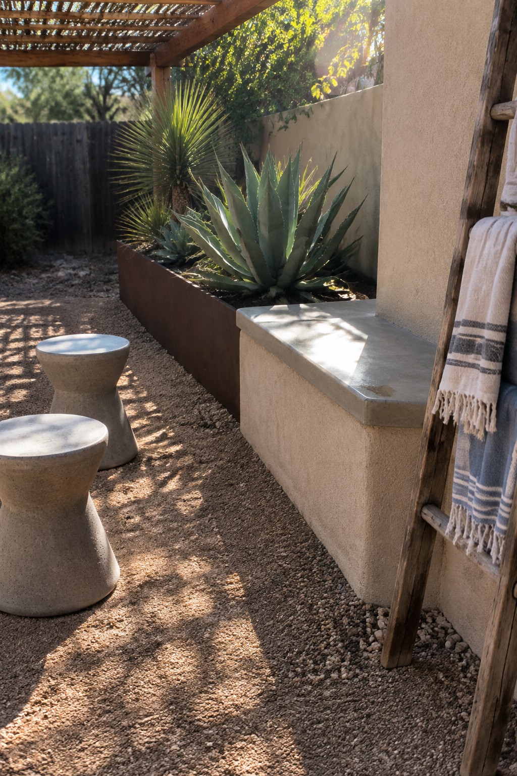 A gravel patio with two concrete stools, a long built-in concrete bench against a stucco wall, a metal planter holding agave plants, and a wooden ladder with towels hanging on it.
