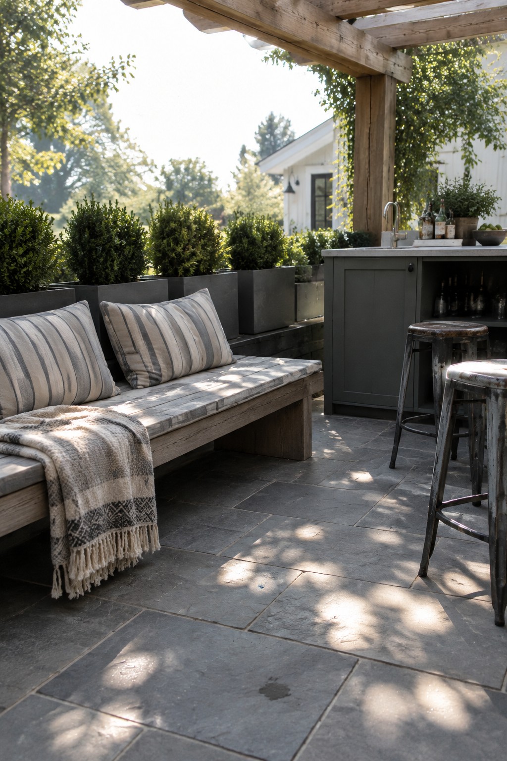 A wooden bench with striped cushions and a throw blanket sits on a stone patio beside a gray outdoor cabinet, metal stools, and large planter boxes under a wooden pergola.