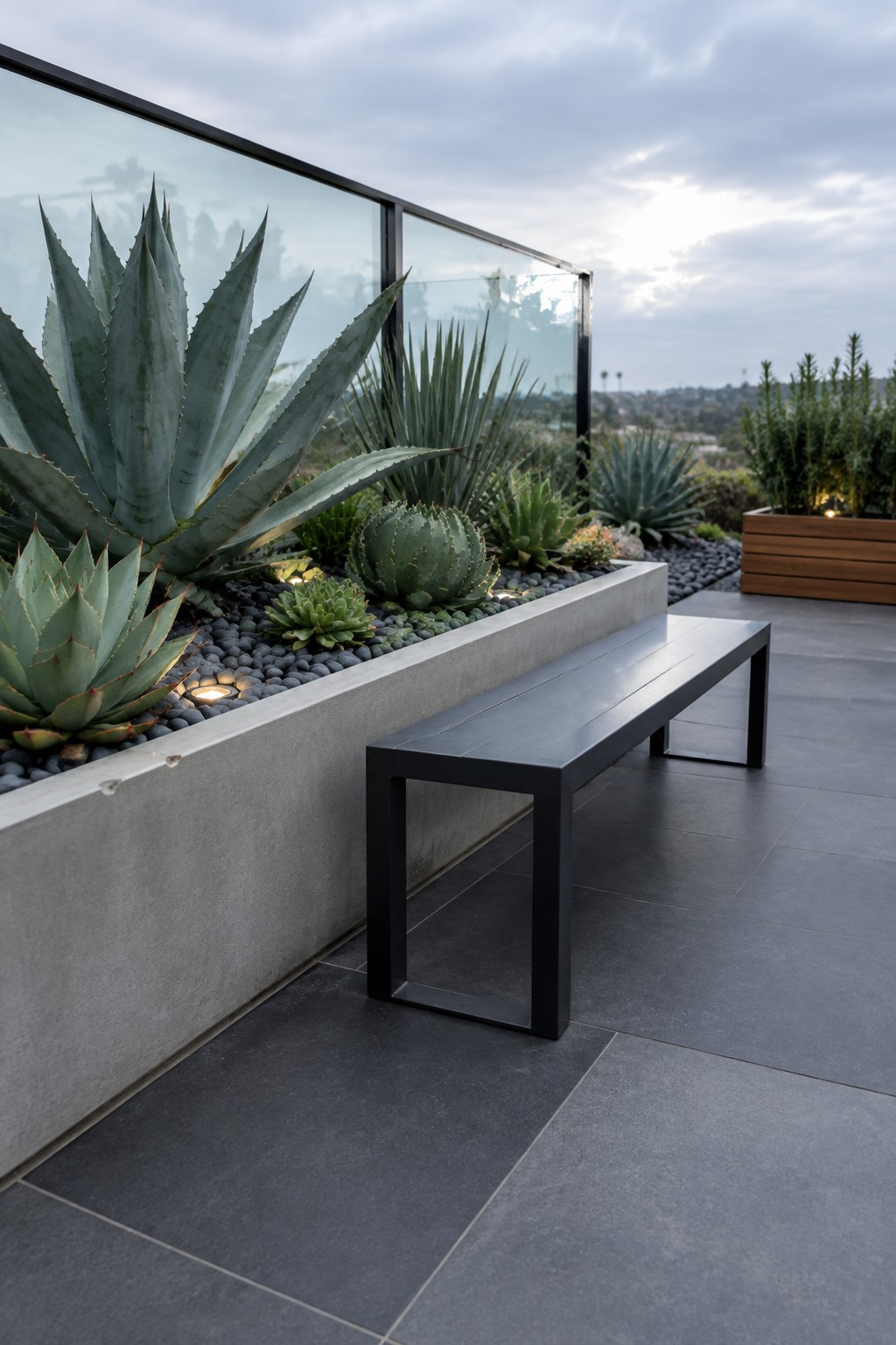 A long concrete planter filled with agaves and succulents runs along a dark-tiled patio beside a black metal bench, with a glass railing and distant hills in view.