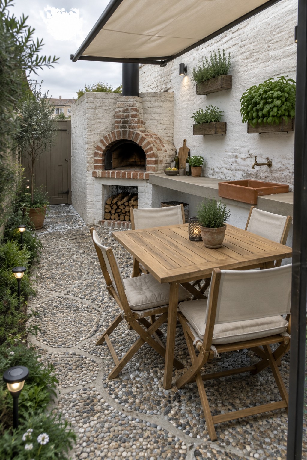 A wooden outdoor dining table with chairs on a cobblestone patio, positioned in front of a brick pizza oven and a concrete counter with a sink against a white wall.