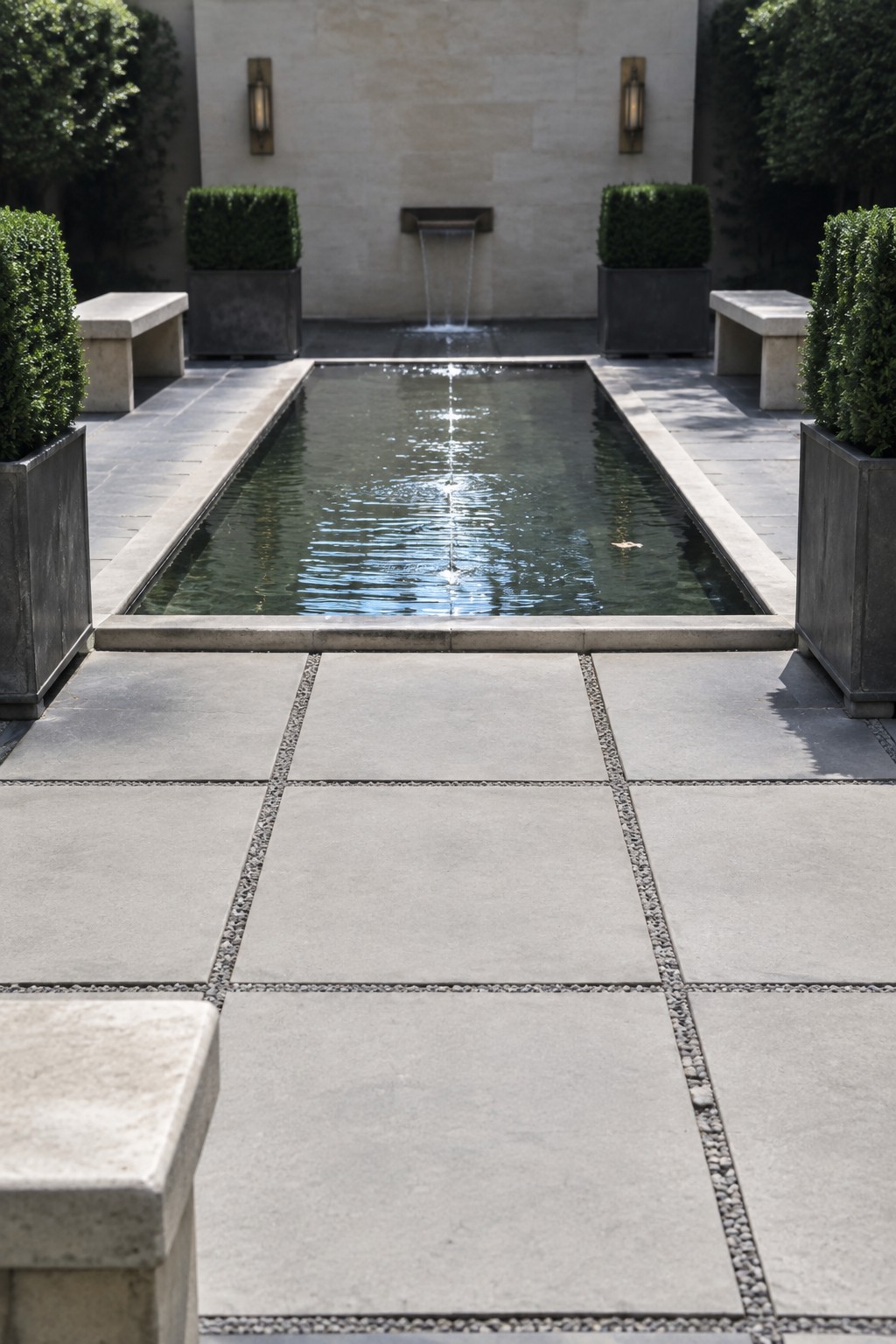 A long rectangular reflecting pool surrounded by large square concrete pavers separated by gravel lines, with potted shrubs and stone benches on either side.