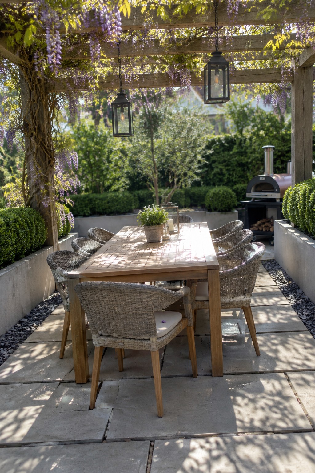 A wooden dining table with woven chairs on large concrete pavers under a pergola covered in wisteria, with lanterns hanging above and a pizza oven visible in the background.