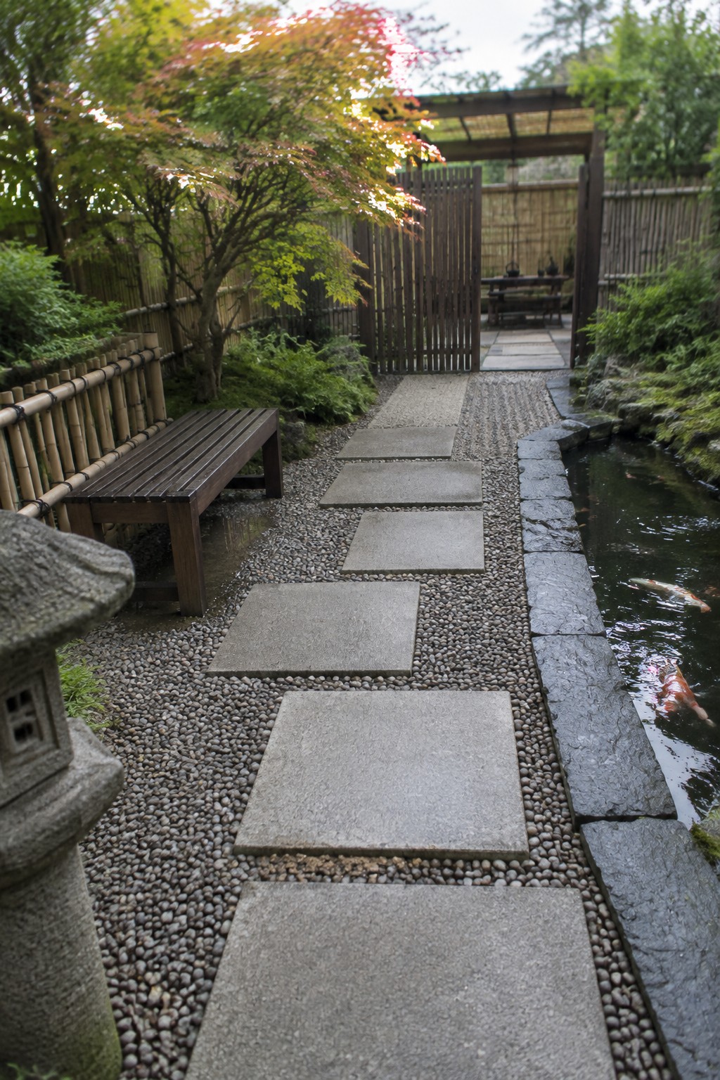 A garden path of large rectangular concrete stepping stones set in gravel runs beside a narrow pond with koi fish, bordered by trees and a bamboo fence.