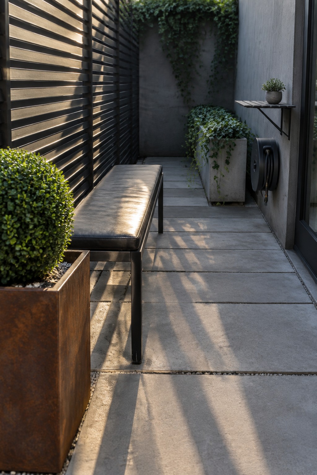 A narrow concrete patio with a long black bench against a dark slatted wall, flanked by a rusted planter and concrete planters with trailing plants.