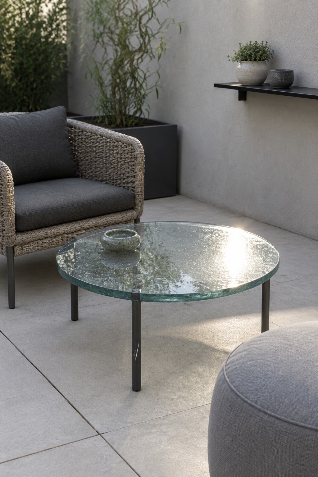 Modern patio with woven gray chair, round glass table, gray ottoman, and potted plants.