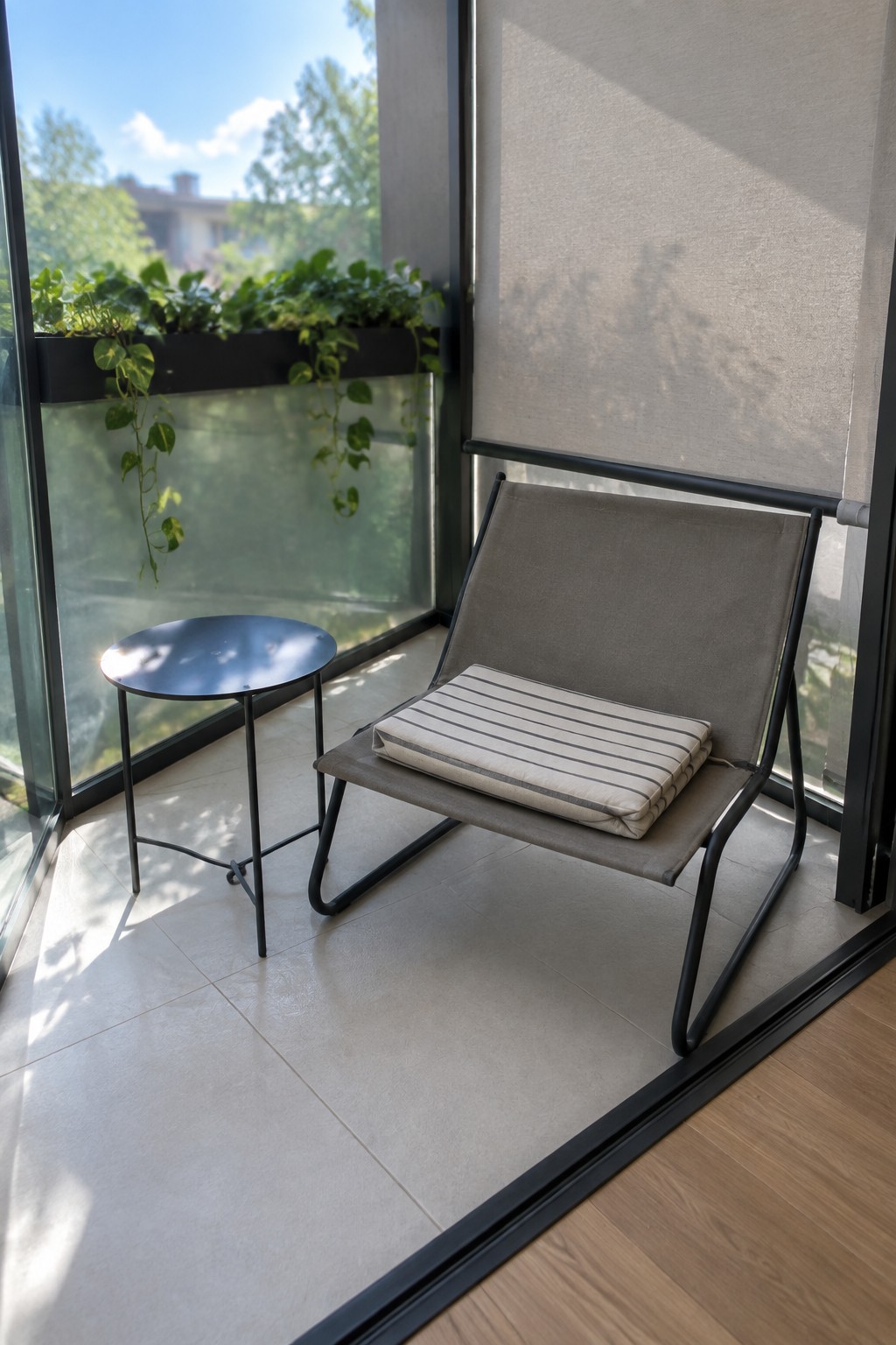 A small balcony with a low gray fabric lounge chair on a black metal frame, a round black side table, and hanging plants along the glass railing.