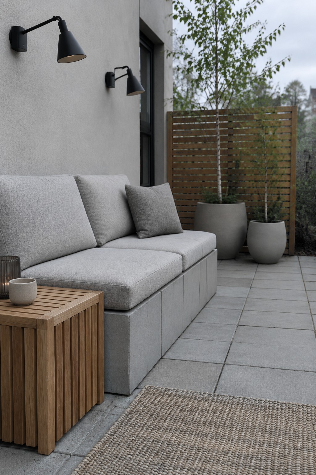 A long bench with gray cushions sits on stacked concrete blocks along a patio wall, next to a wooden slatted side table and black wall lights.