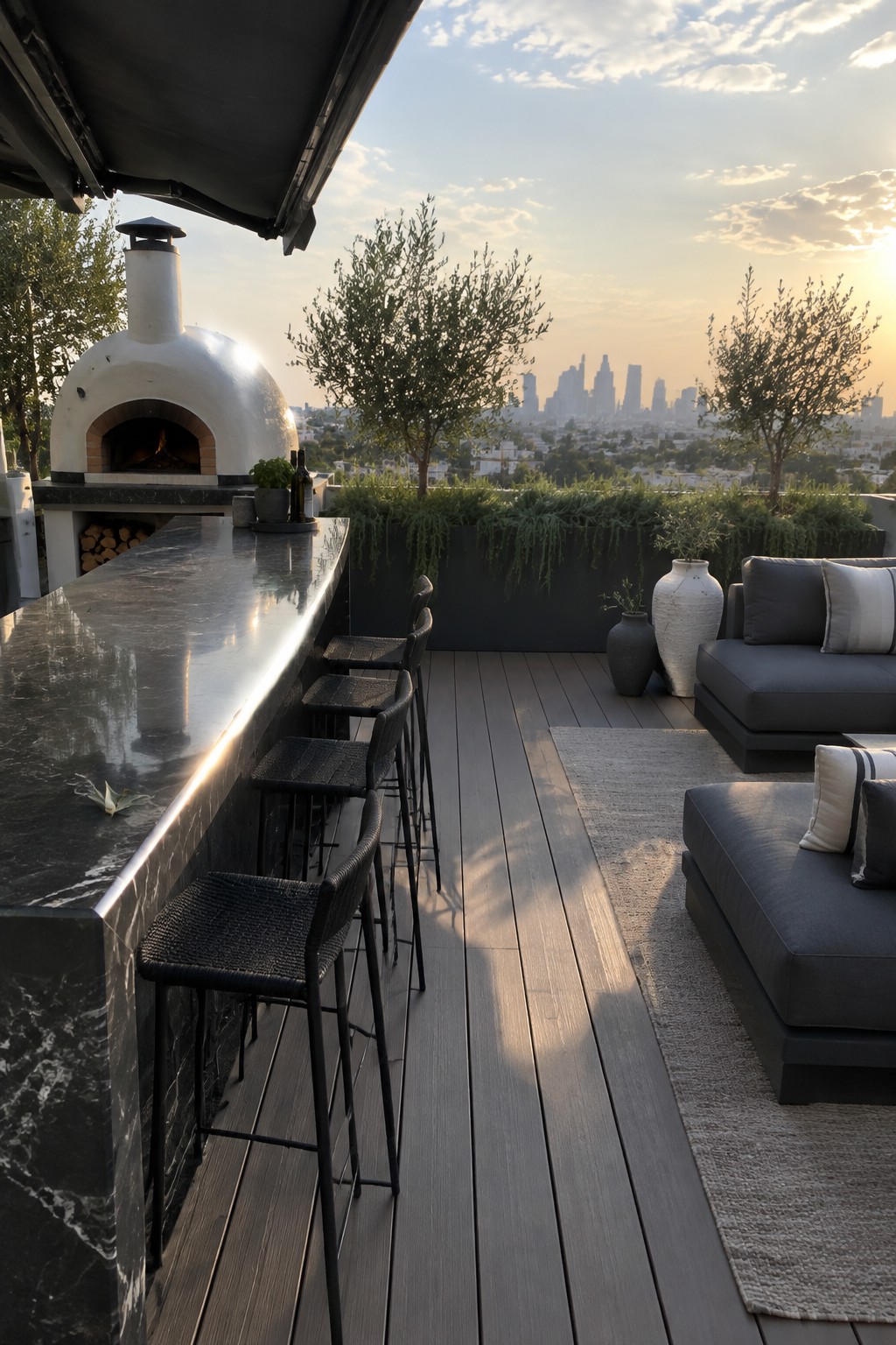 A modern rooftop patio with a white pizza oven, long dark marble bar counter, black woven bar stools, gray outdoor sofas on a rug, and a city skyline view at sunset.