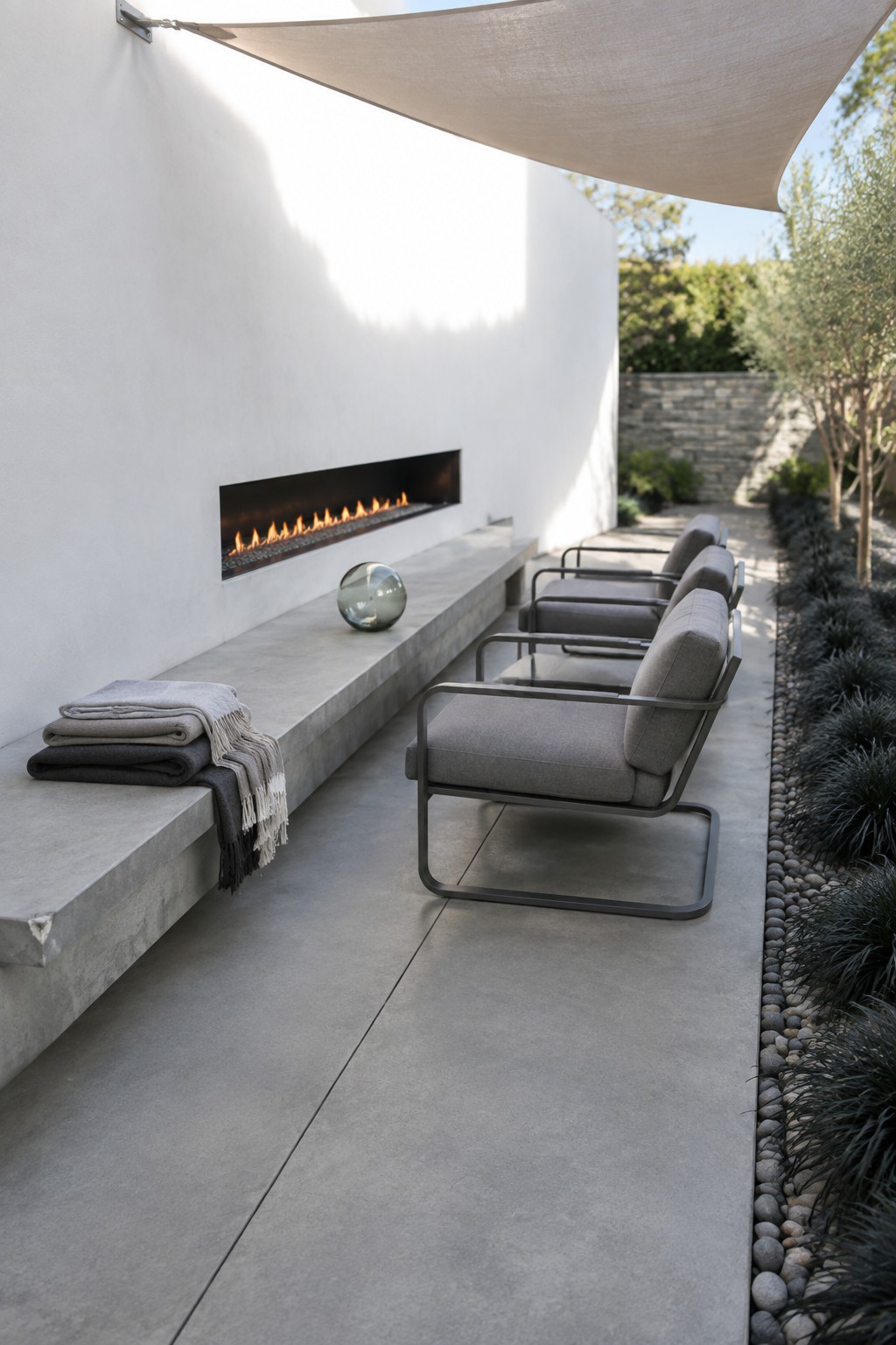 Minimalist patio with linear fireplace, gray armchairs, concrete bench, folded throws, and shade sail.
