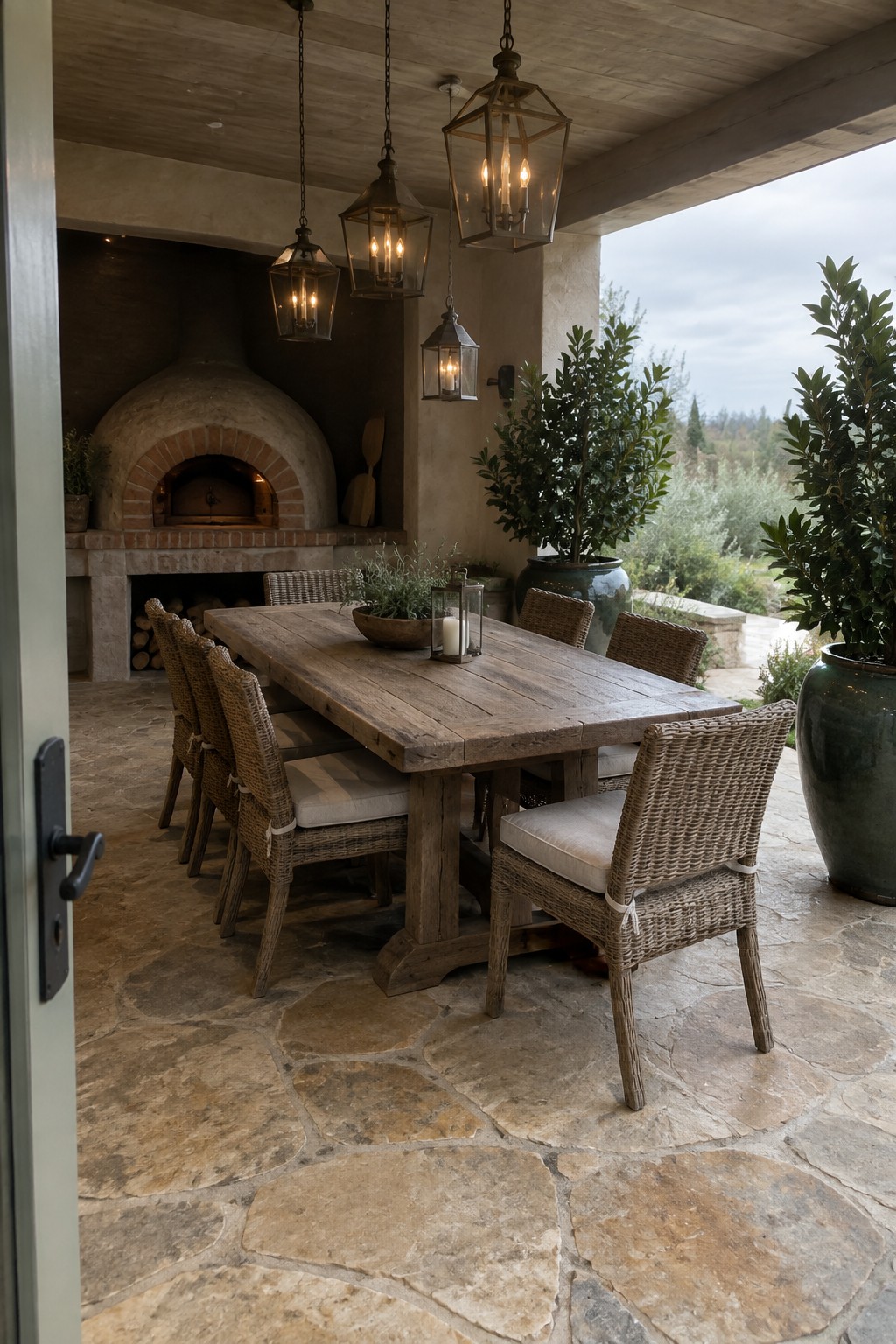 A long wooden dining table with woven chairs on a flagstone patio, positioned near a brick oven and large potted plants under hanging lanterns.