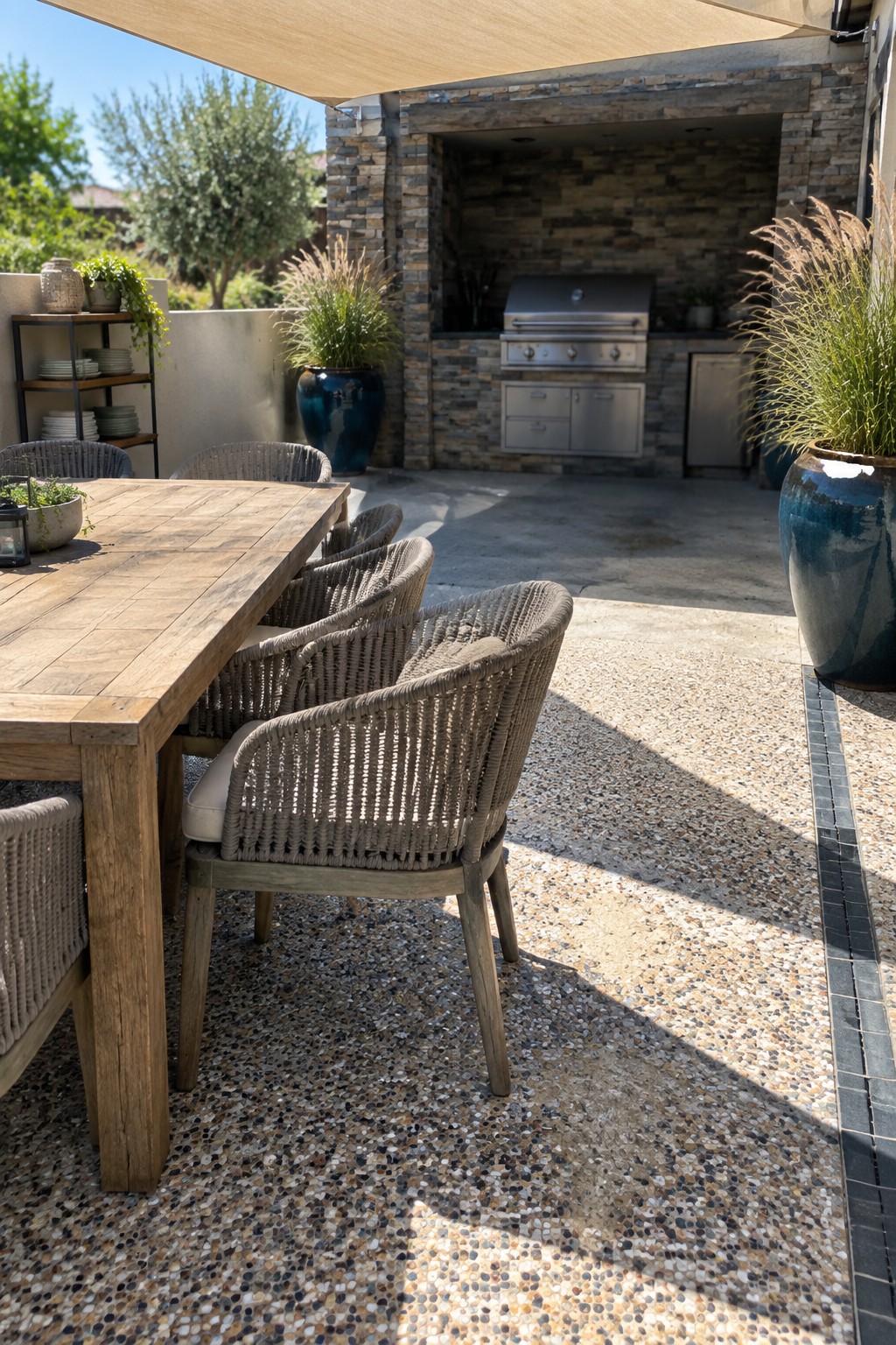 An outdoor patio dining area with a wooden table, woven chairs, and speckled pebble flooring in front of a stone outdoor kitchen.