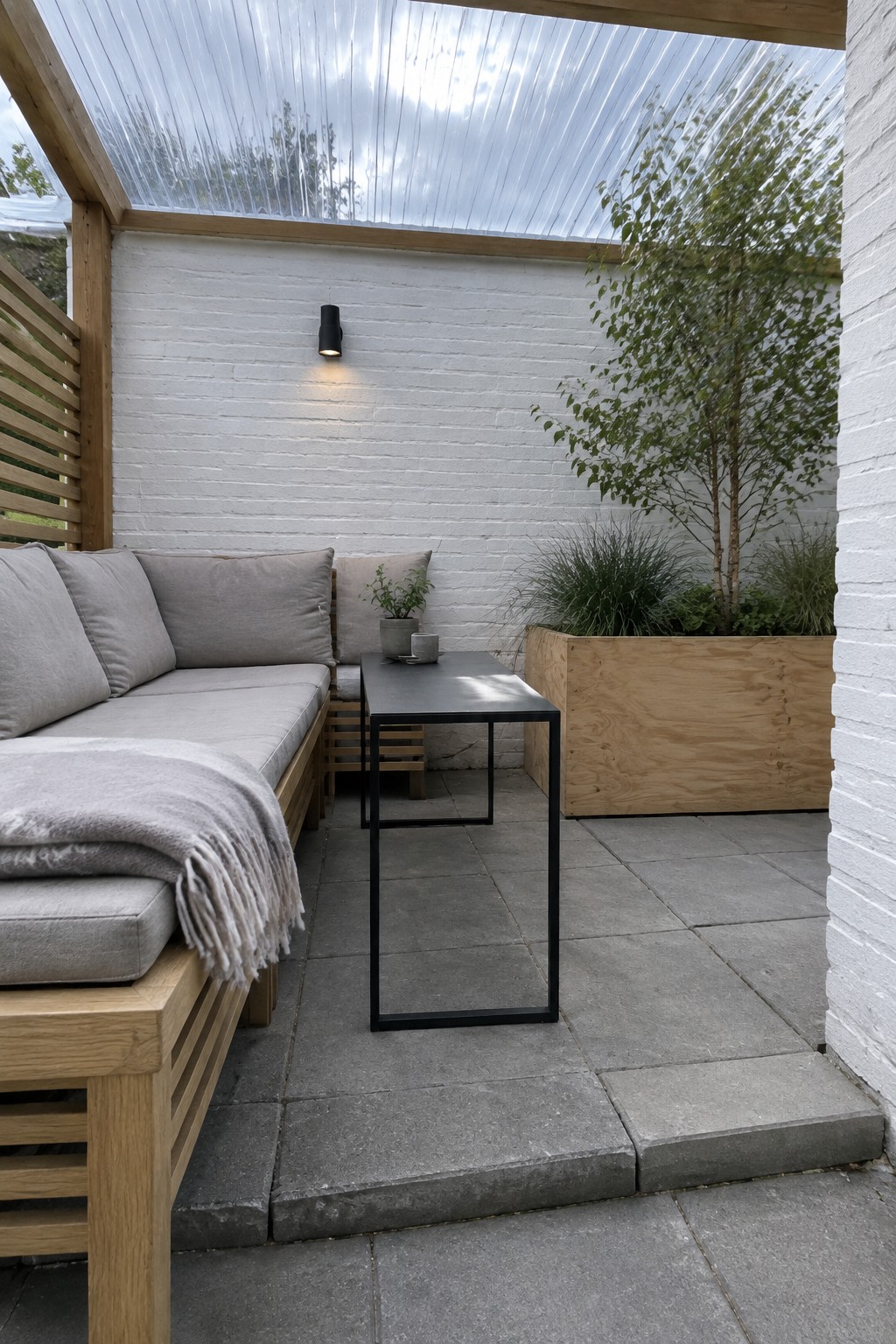 A covered courtyard patio with a translucent roof, a cushioned wood sofa, a narrow black table, and a raised wooden planter.