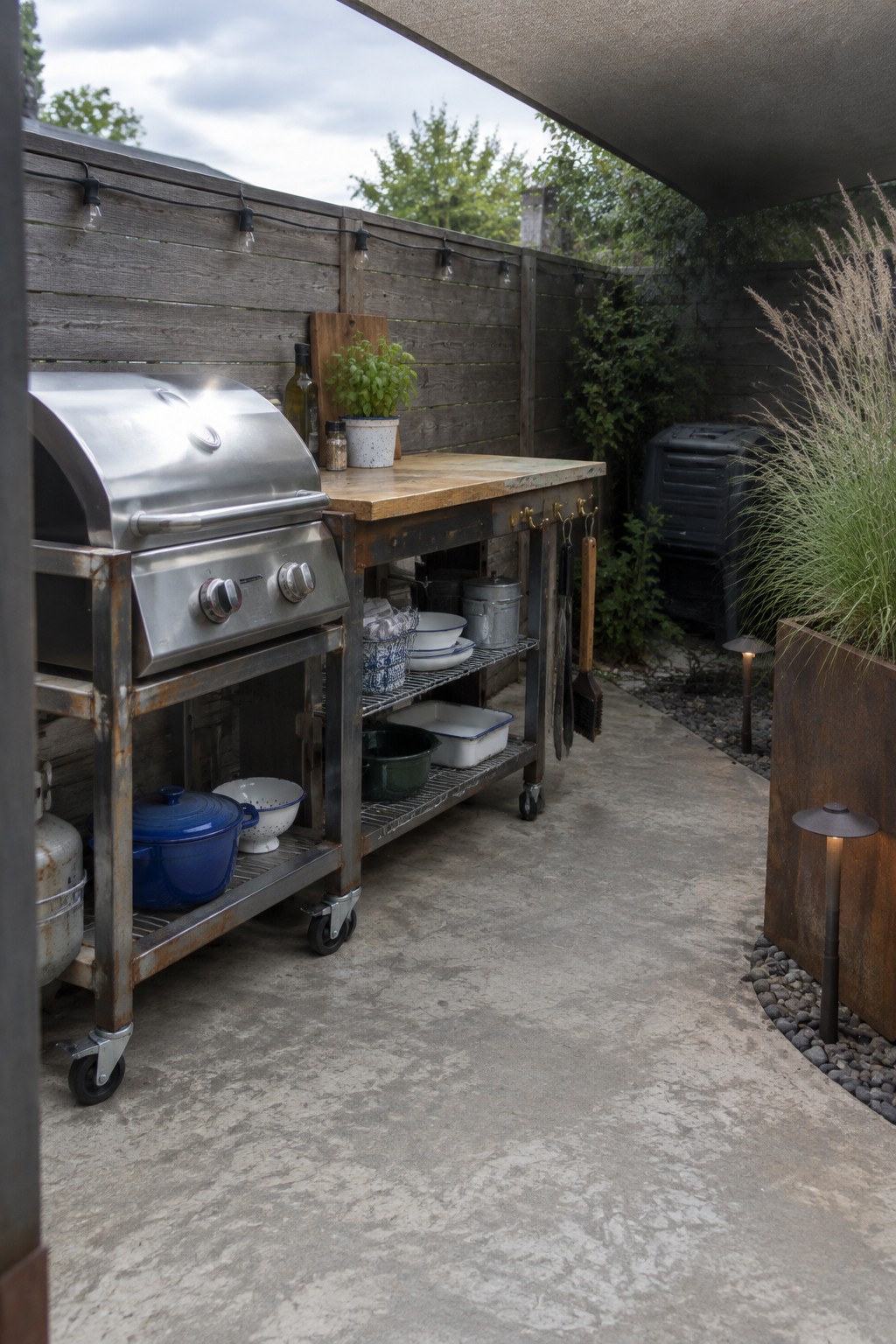 A stainless steel gas grill stands beside a wooden prep counter with shelves in a narrow patio.