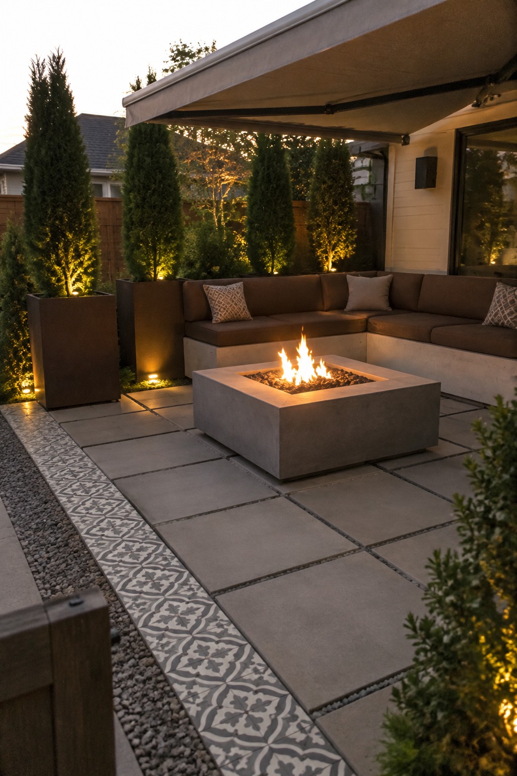 A paved patio with tall evergreen plants in rectangular planters, an L-shaped sofa, and a concrete fire table beneath an awning.